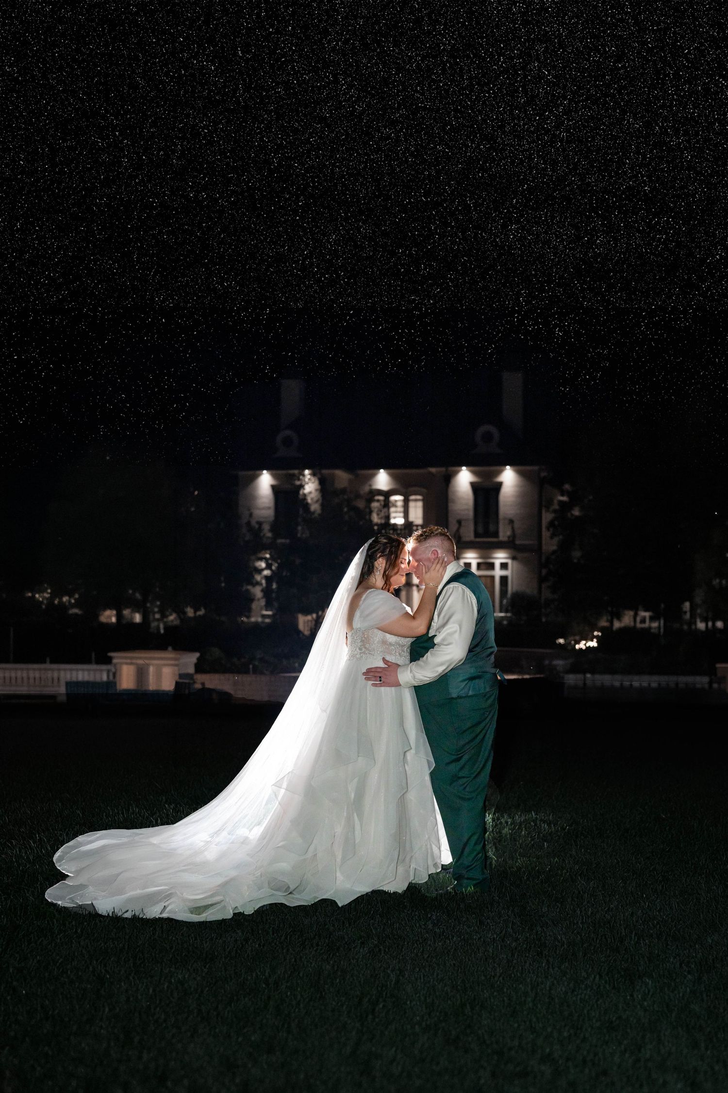 Bride in a long white gown and veil kisses her groom in a green vest on a lawn at night, with an illuminated manor behind them.