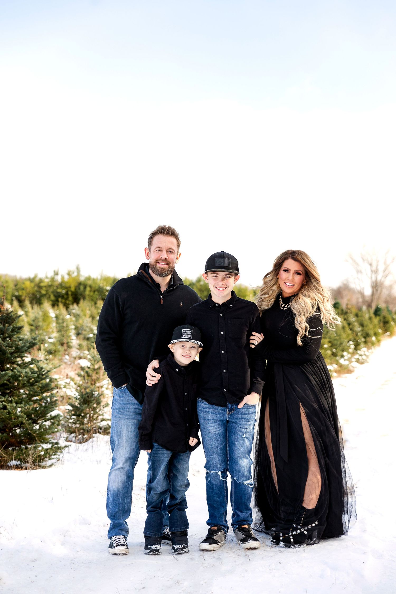 A smiling family of four in black outfits poses together in the snow at an evergreen Christmas tree farm on a bright winter day.