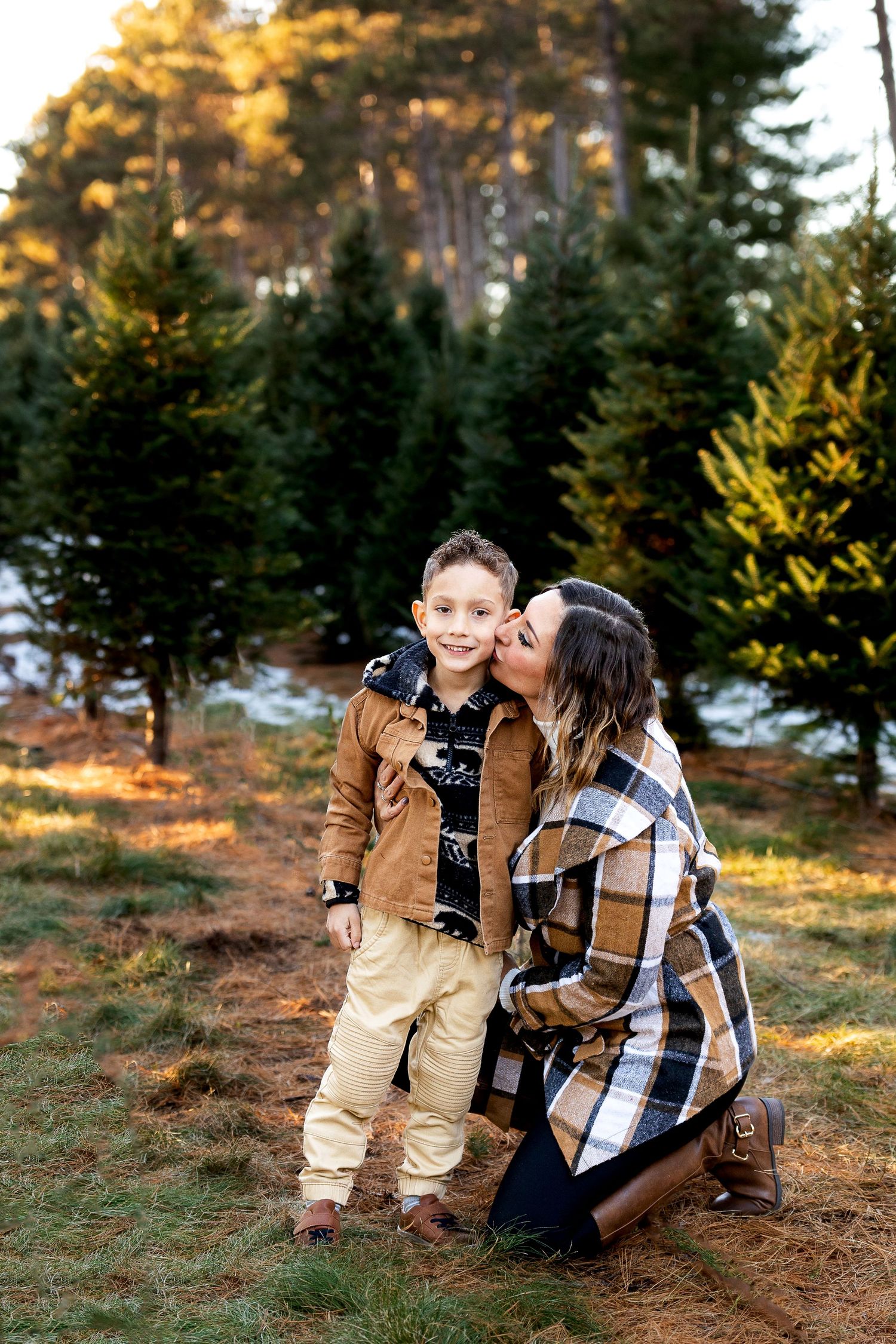 A smiling boy in a tan jacket is kissed on the cheek by his kneeling mother in a plaid coat at an evergreen Christmas tree farm.
