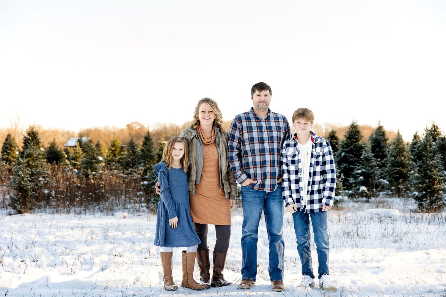 Family of four in plaid shirts and boots posing together on a snowy Christmas tree farm with evergreen trees in the background.
