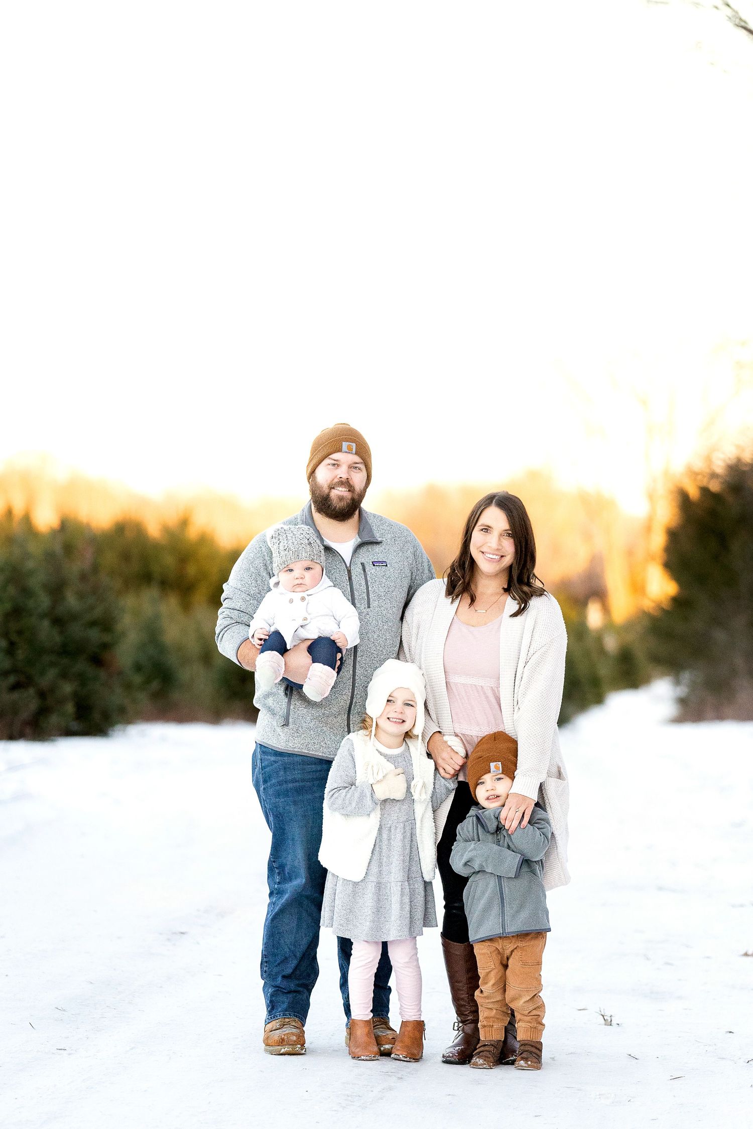 Family of five in cozy winter outfits and beanies posing together on a snowy path with evergreen trees in the background.