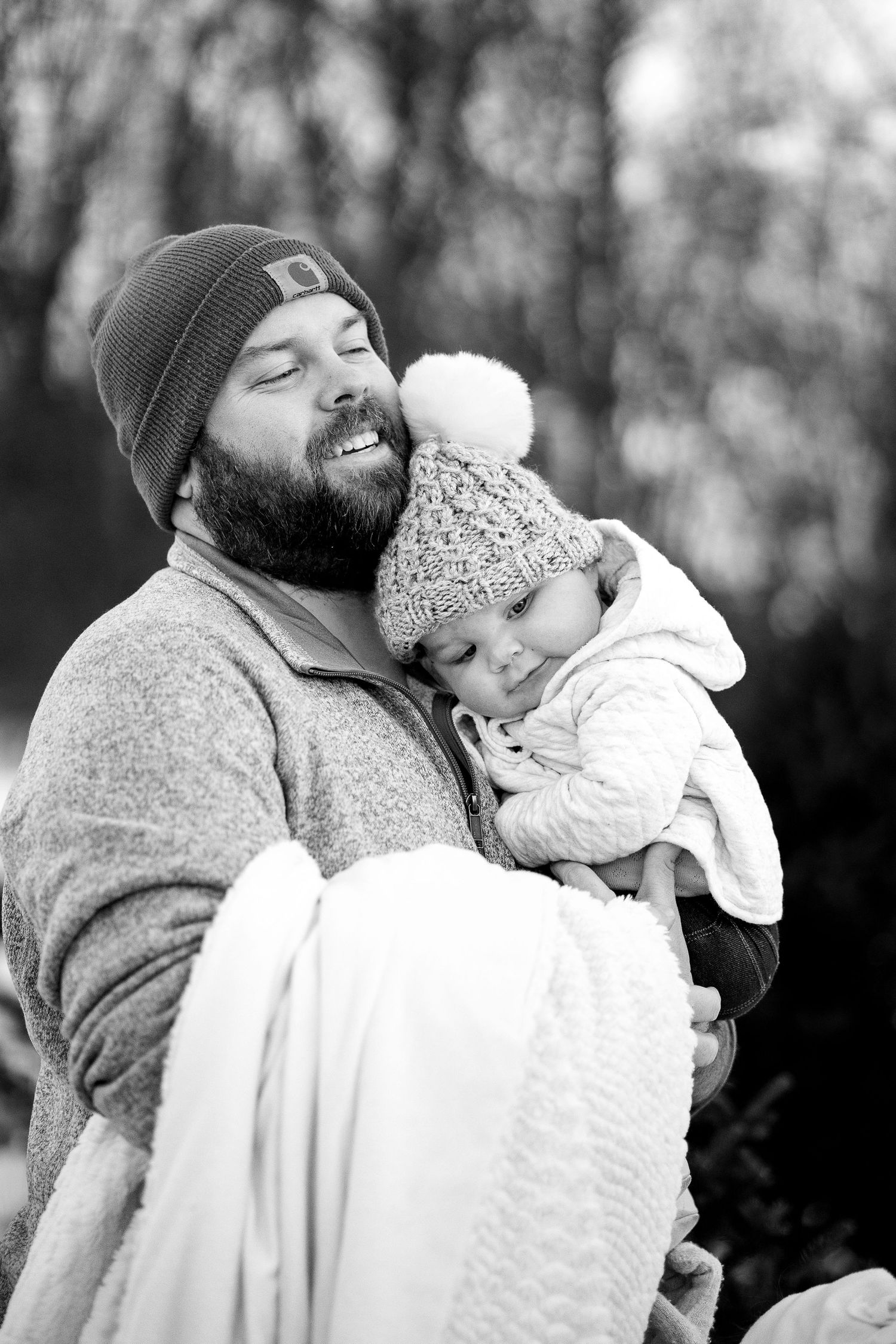 Bearded man in a beanie smiles with eyes closed while holding a sleepy baby in a pom-pom knit hat, black and white winter portrait.