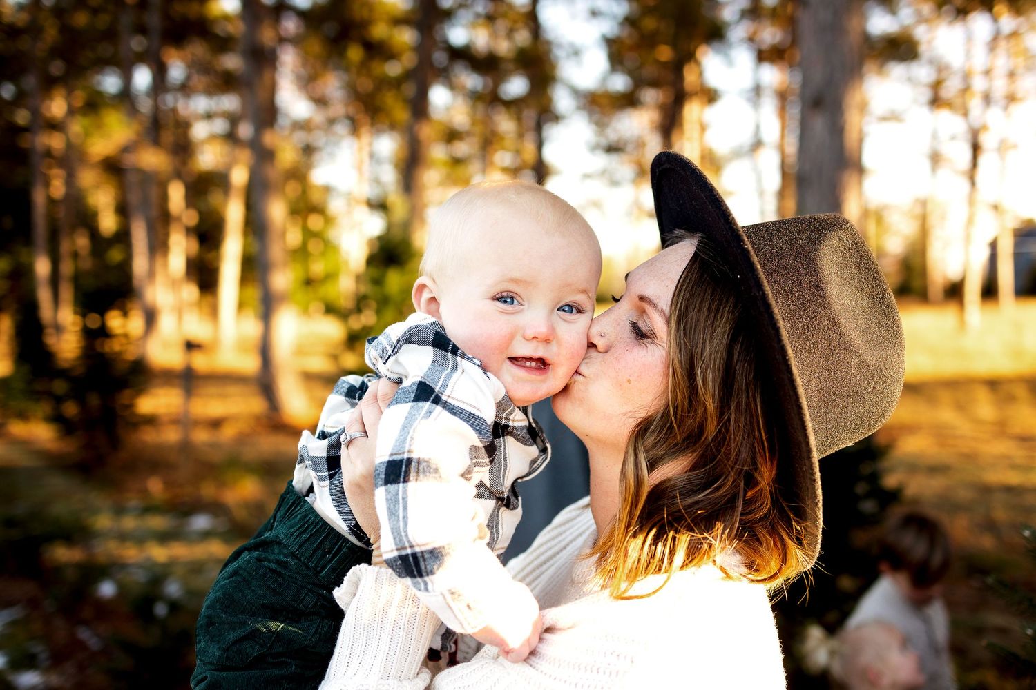 Woman in a brown felt hat kisses a smiling baby in a plaid shirt while standing outdoors among tall trees in warm golden light.