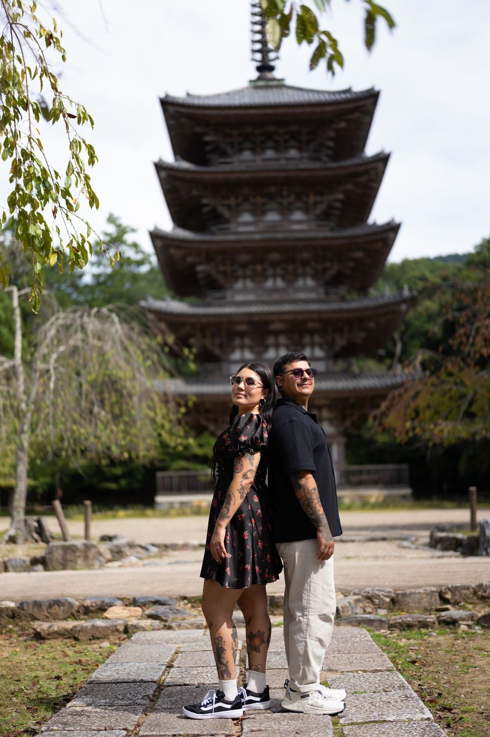 Tattooed couple in sunglasses stand back to back on a stone path before a multi-tiered Japanese pagoda surrounded by green trees.