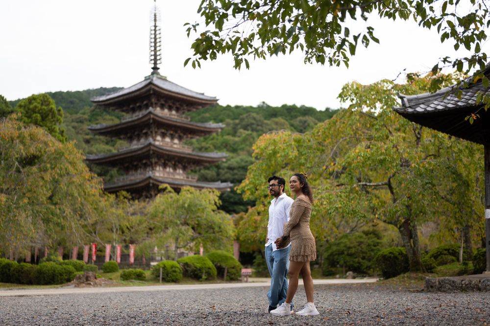 Couple walking hand in hand on a gravel path in a Japanese garden with a tall multi-tiered pagoda and lush green trees behind them.