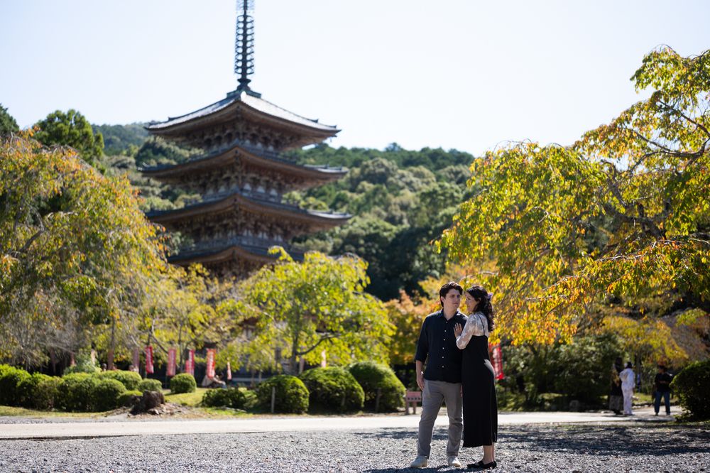Couple embracing in front of a multi-tiered Japanese pagoda surrounded by golden autumn foliage and green hills on a sunny day.