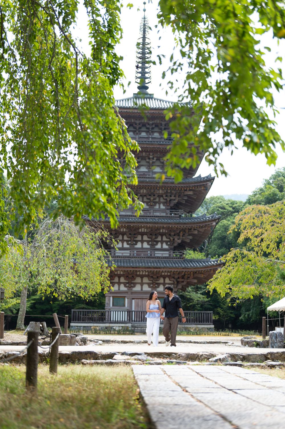 Couple walking along a stone path toward a historic multi-tiered Japanese pagoda surrounded by lush green trees.