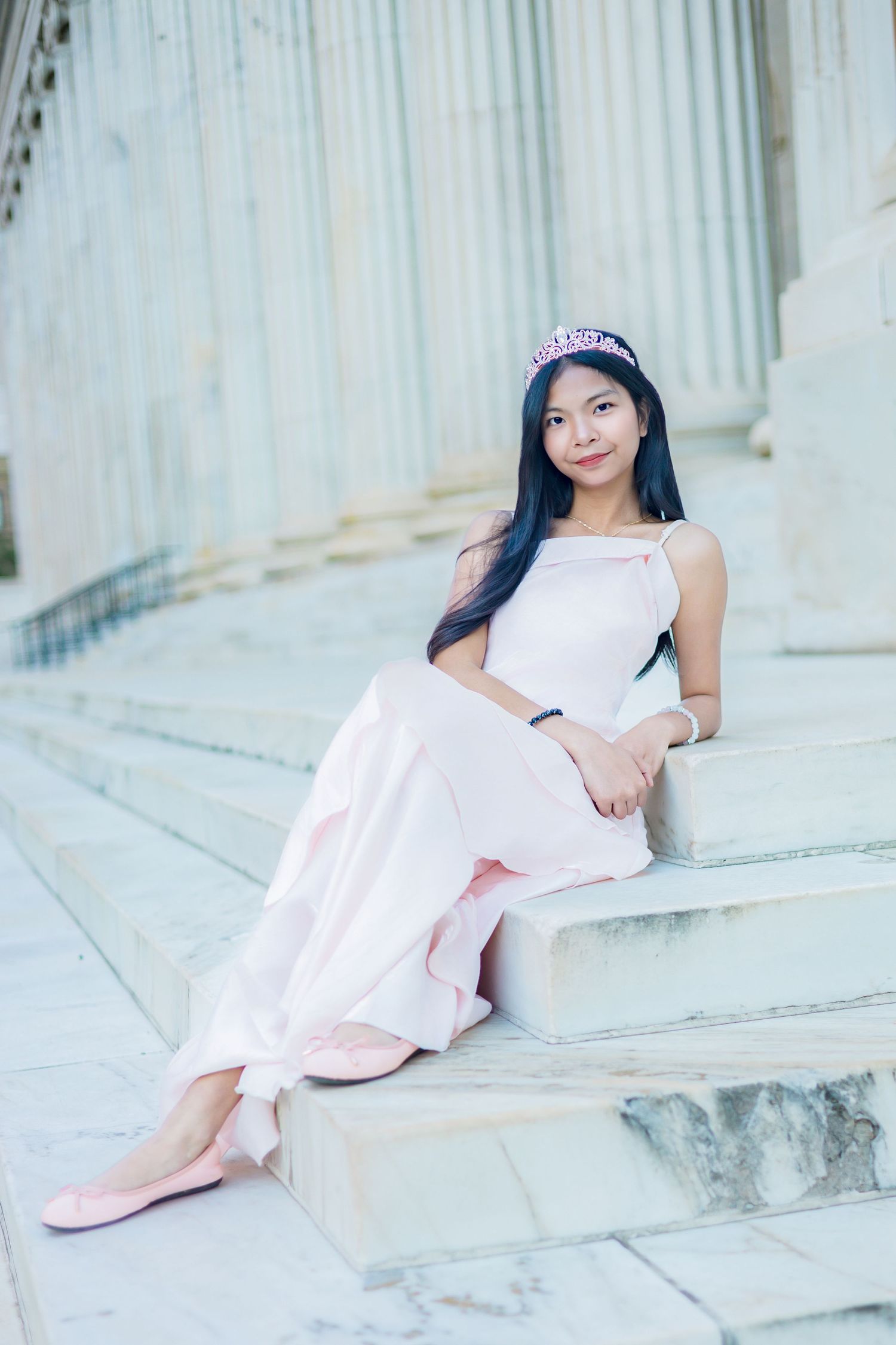 Young woman in pink dress and tiara sits gracefully on marble steps beside classical columns.