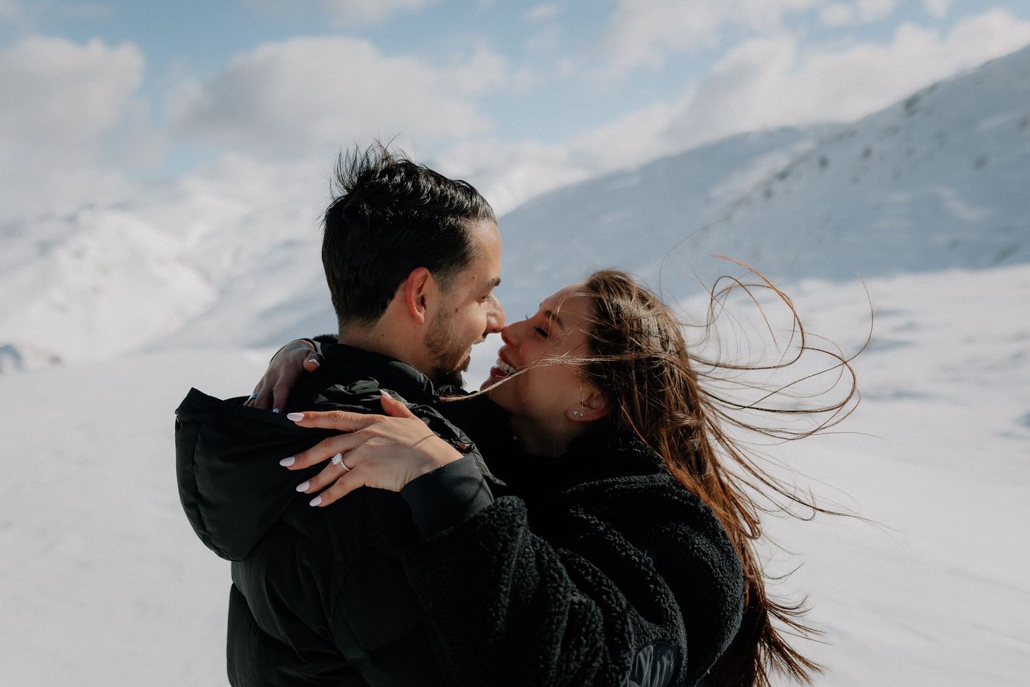 Couple embracing and smiling at each other on a snowy mountain, woman's hair blowing in the wind.
