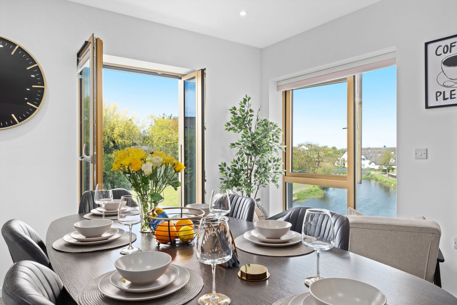 Modern dining room with round table set for six, yellow flowers, and large windows overlooking a scenic waterway.