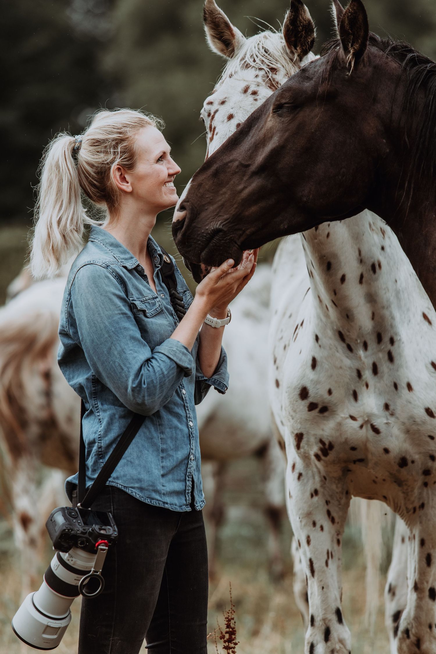 Blonde woman in denim shirt holding camera interacts with a spotted Appaloosa horse outdoors.