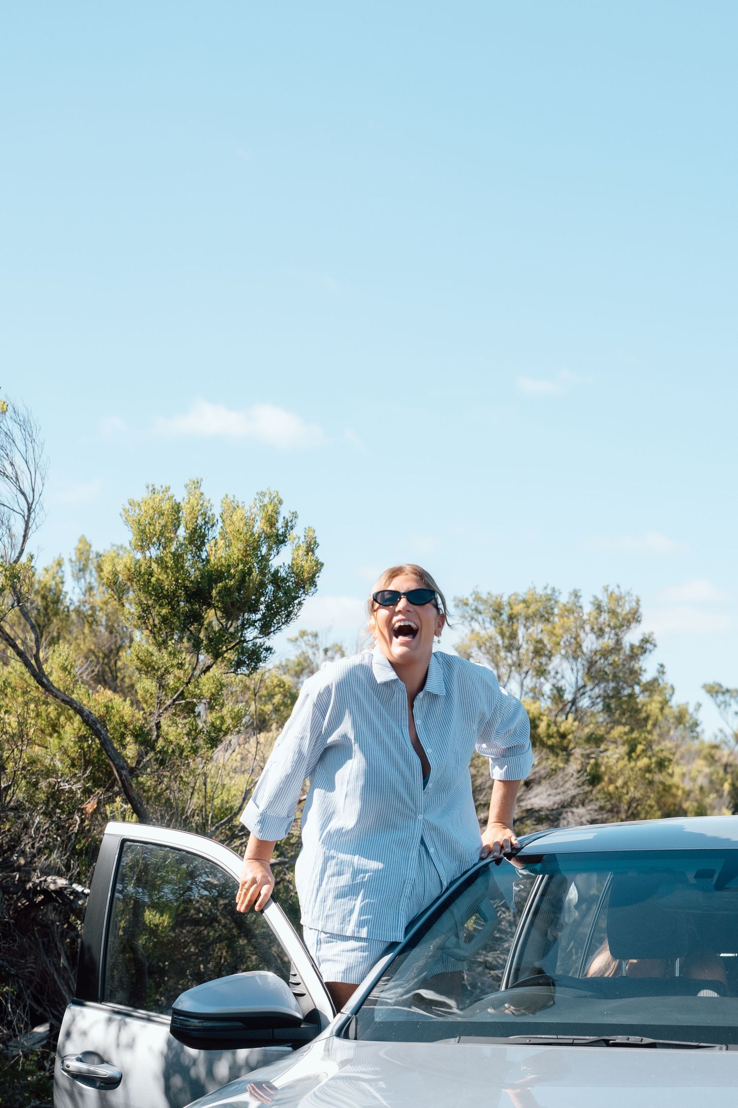 Happy senior woman in sunglasses standing by white car outdoors on sunny day with blue sky and green trees.