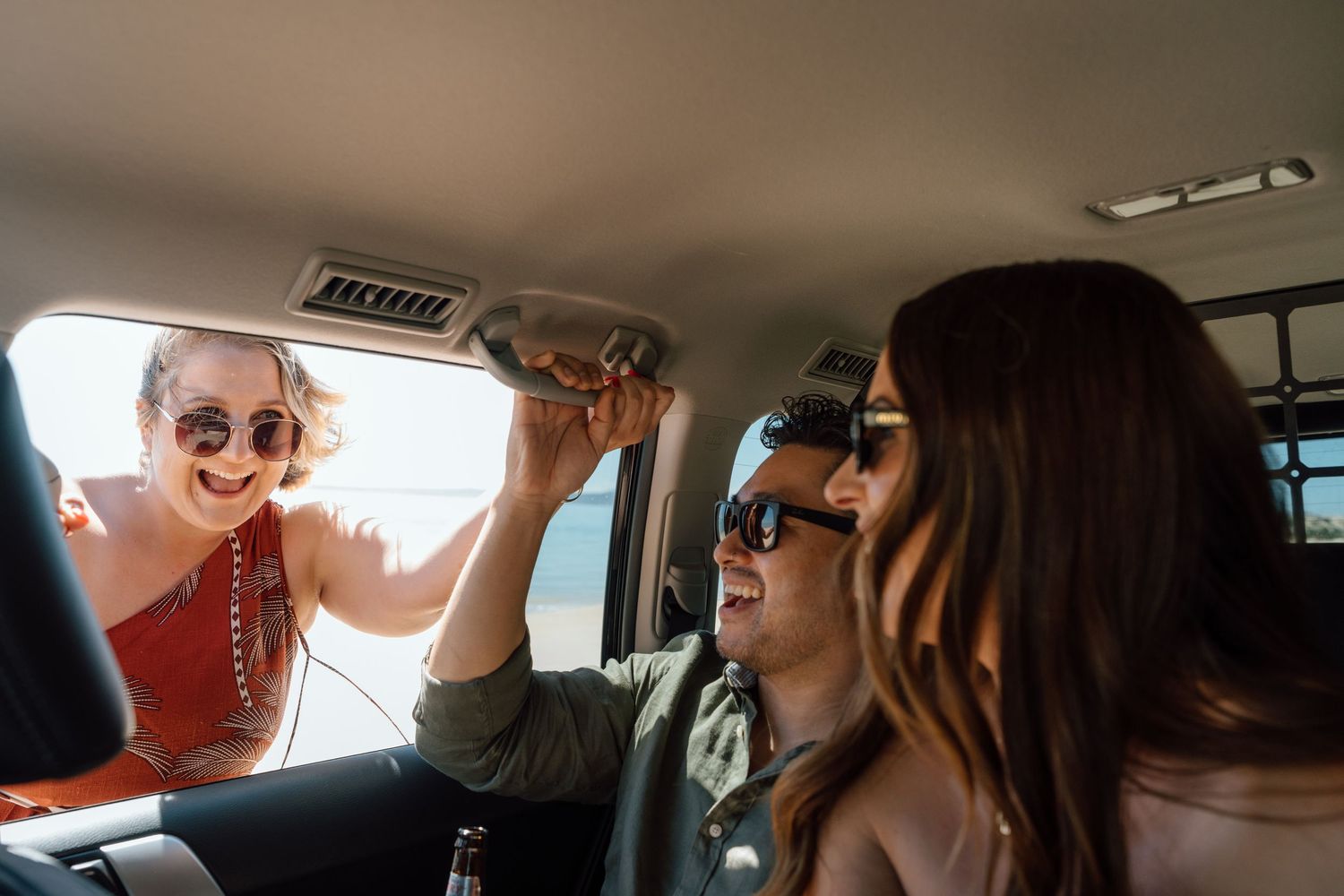 Three friends laughing and having fun on a road trip inside a car, wearing sunglasses on a sunny day.