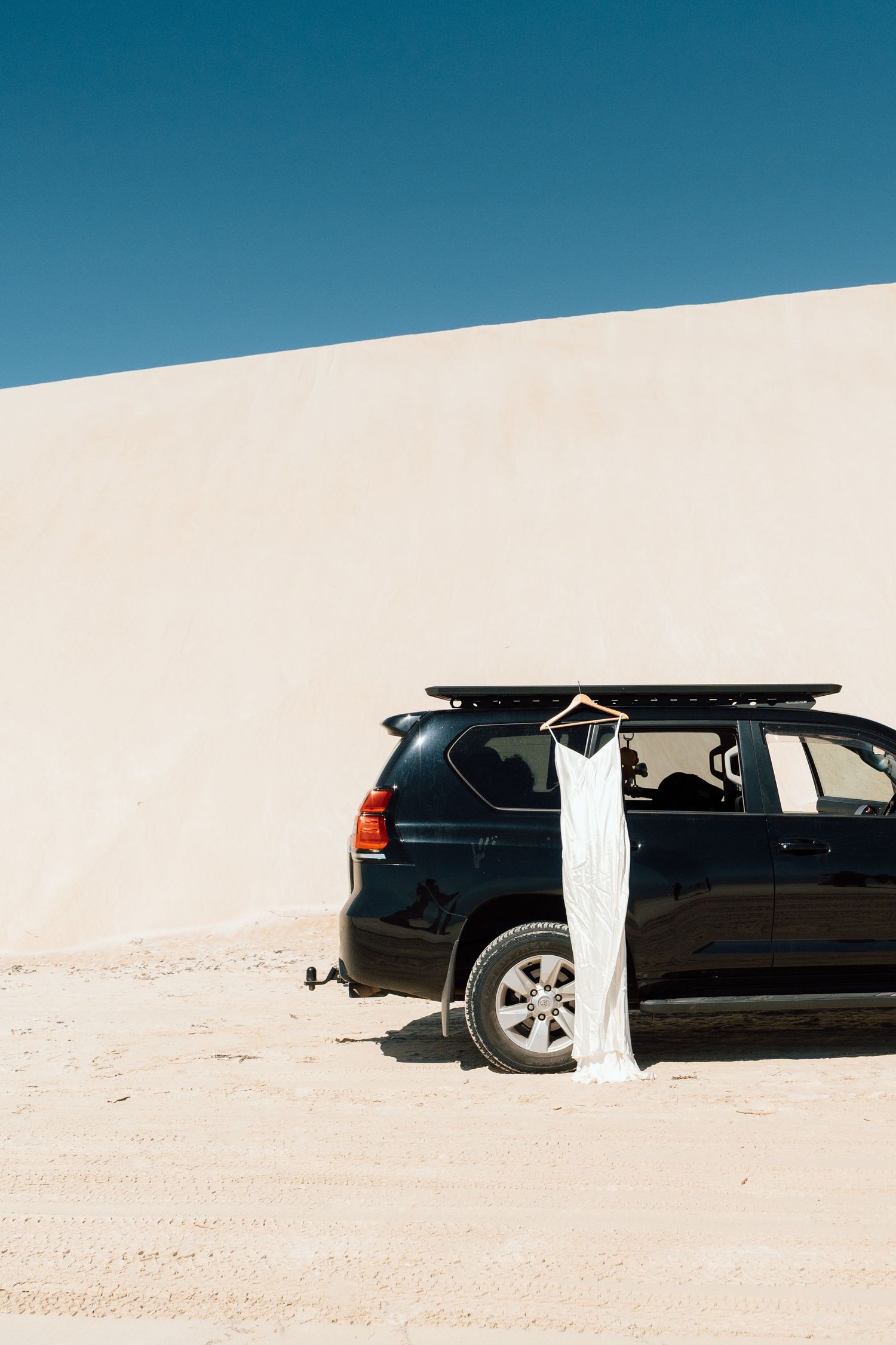 White wedding dress hanging on a black SUV roof rack in a sandy desert landscape under a clear blue sky.