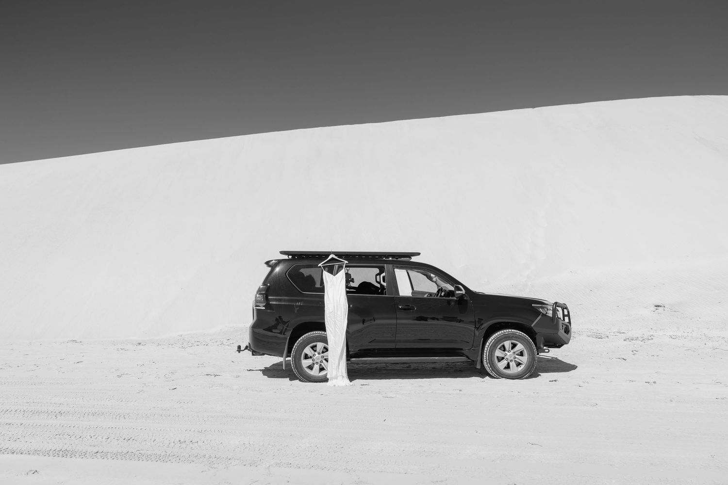 Black and white photo of a person leaning against a dark SUV on white sand dunes in a desert landscape.