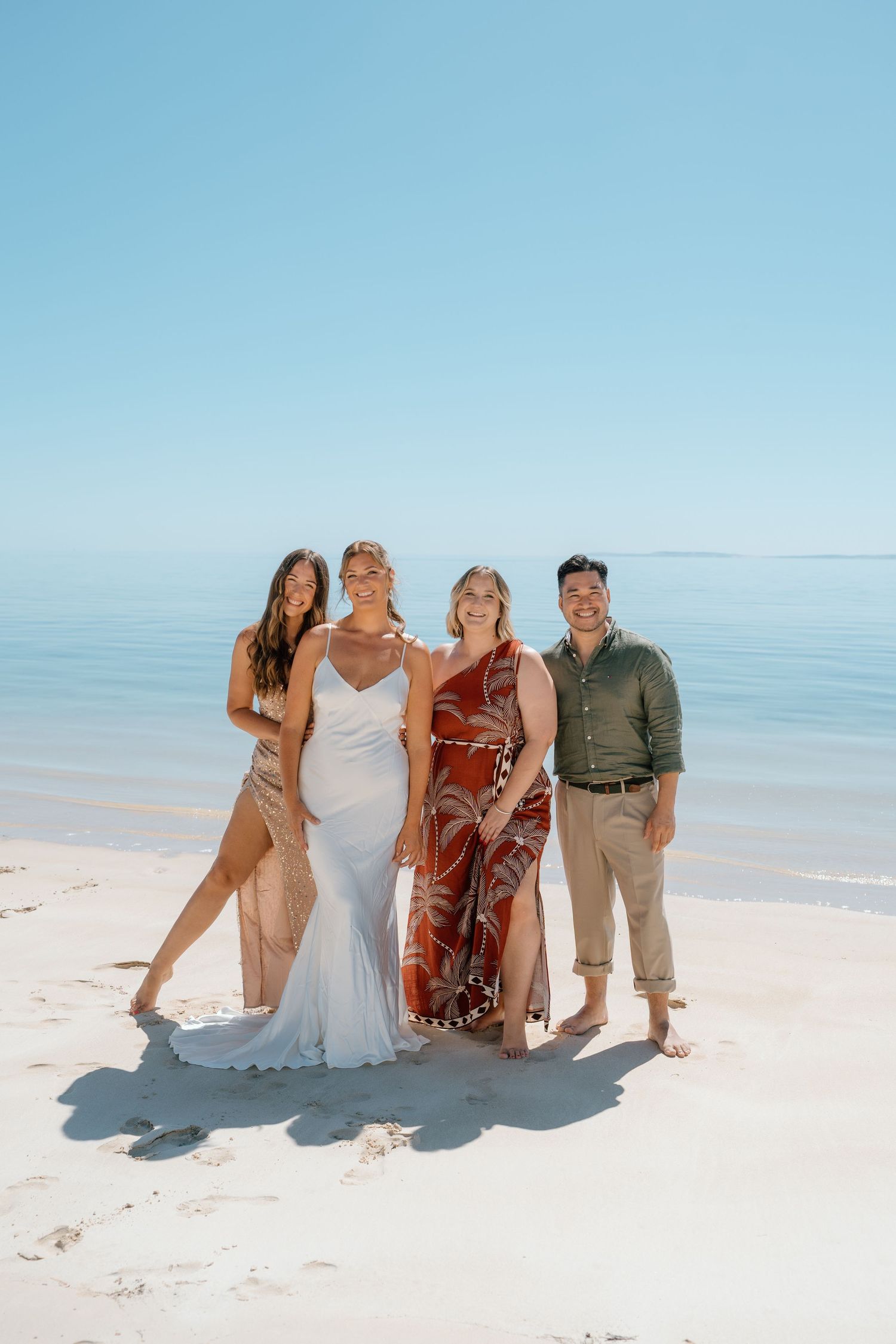 Four people posing together on a white sandy beach, including a bride in a white gown, on a sunny clear day.