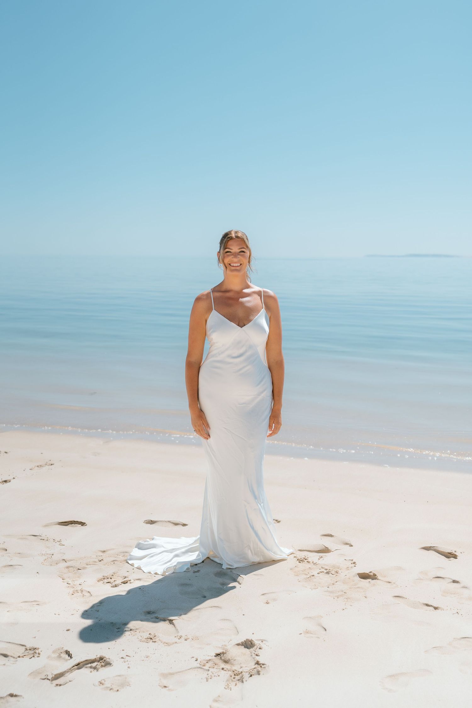 Radiant bride in elegant white satin slip gown standing on pristine white sand beach with clear blue ocean.