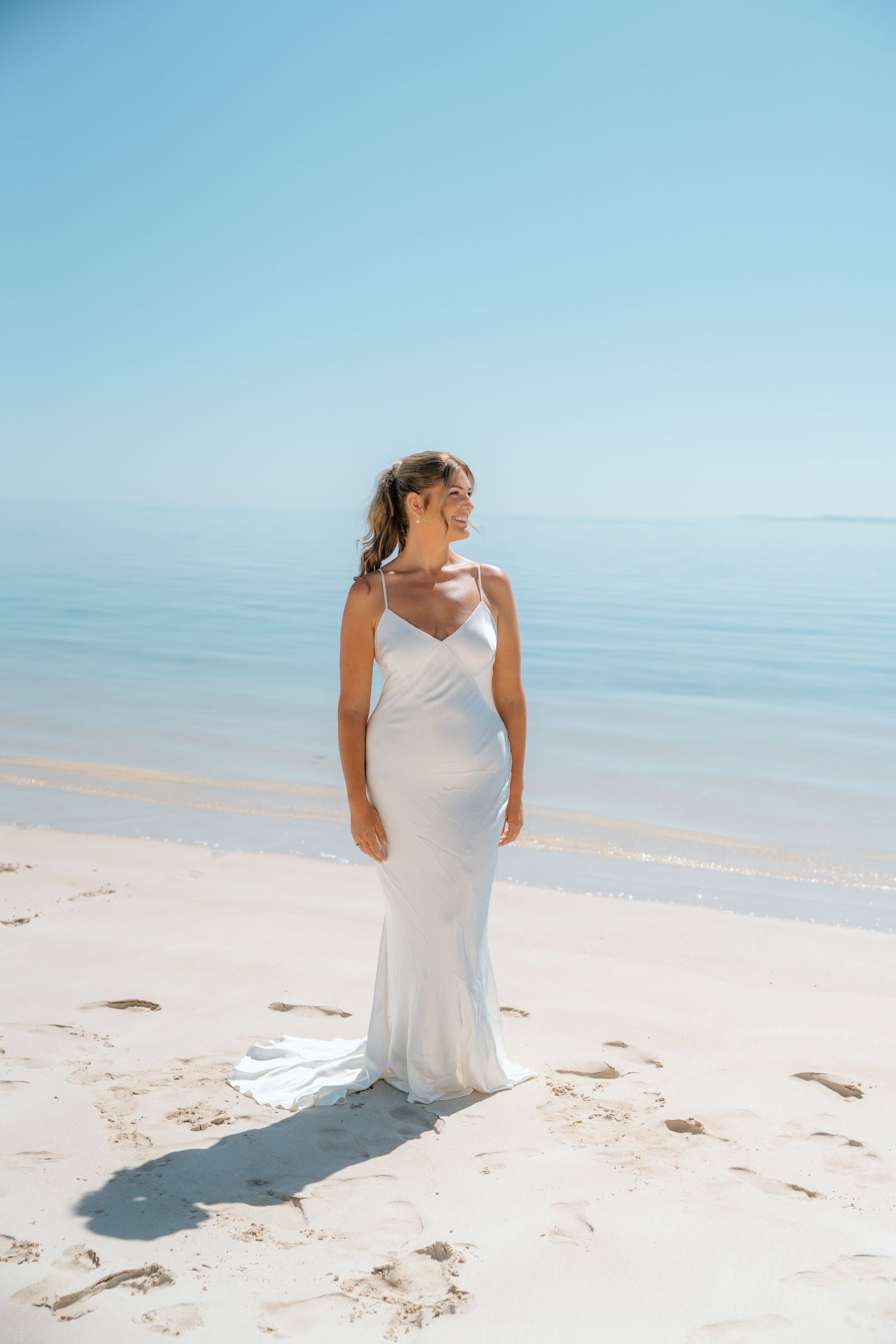 Bride in white satin gown standing on white sand beach with blue ocean backdrop.