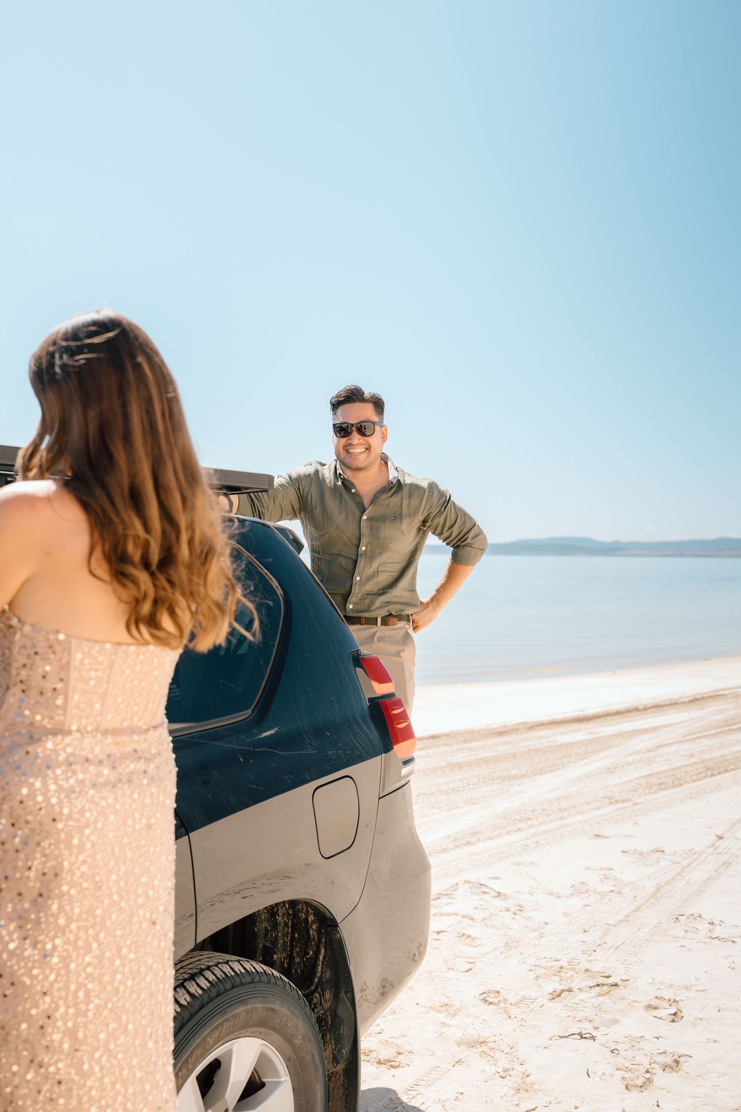 Couple near SUV on sandy beach, man in sunglasses smiling at woman in sequin dress.