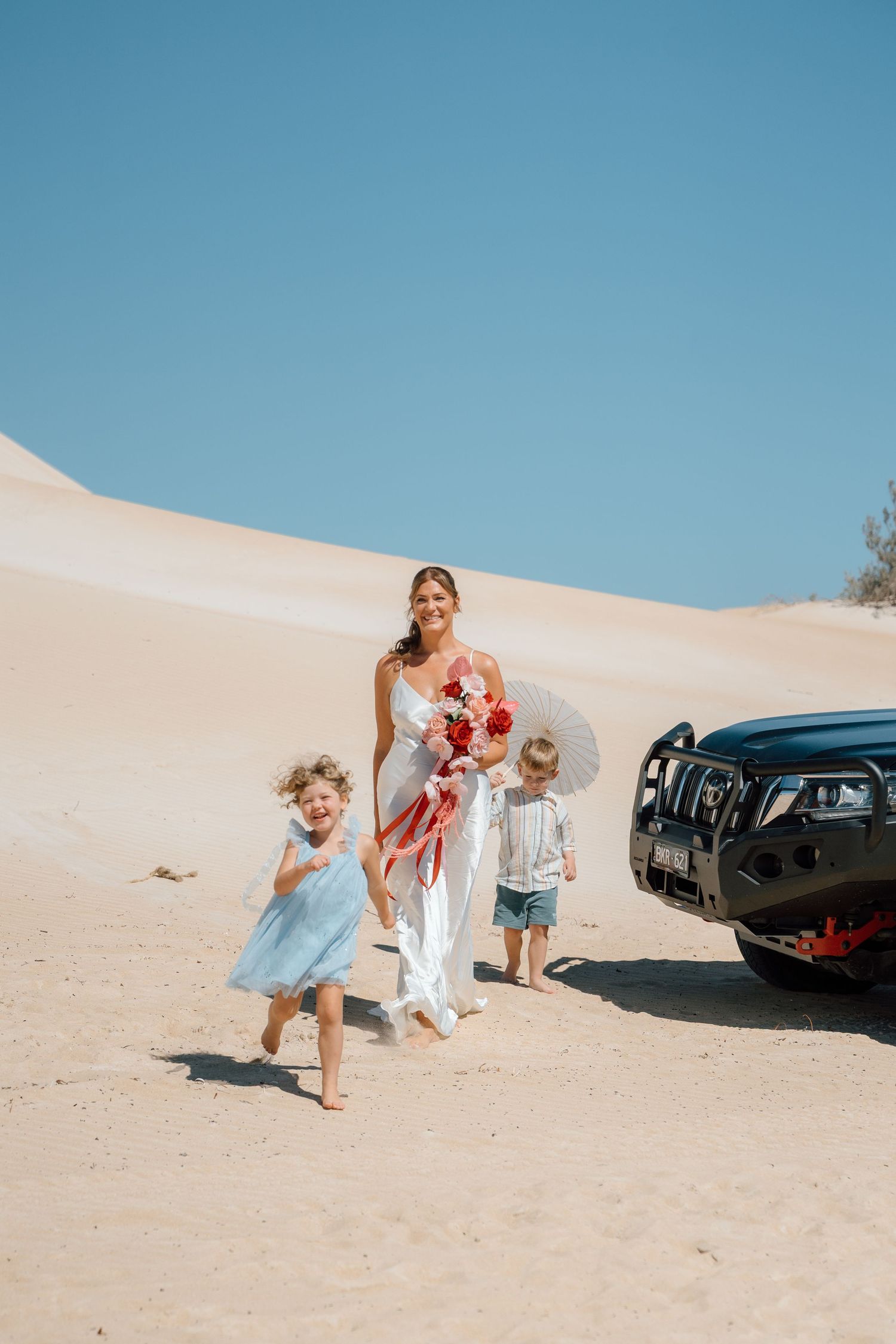 Bride in white dress walking with two young children on sandy beach beside a black 4WD.