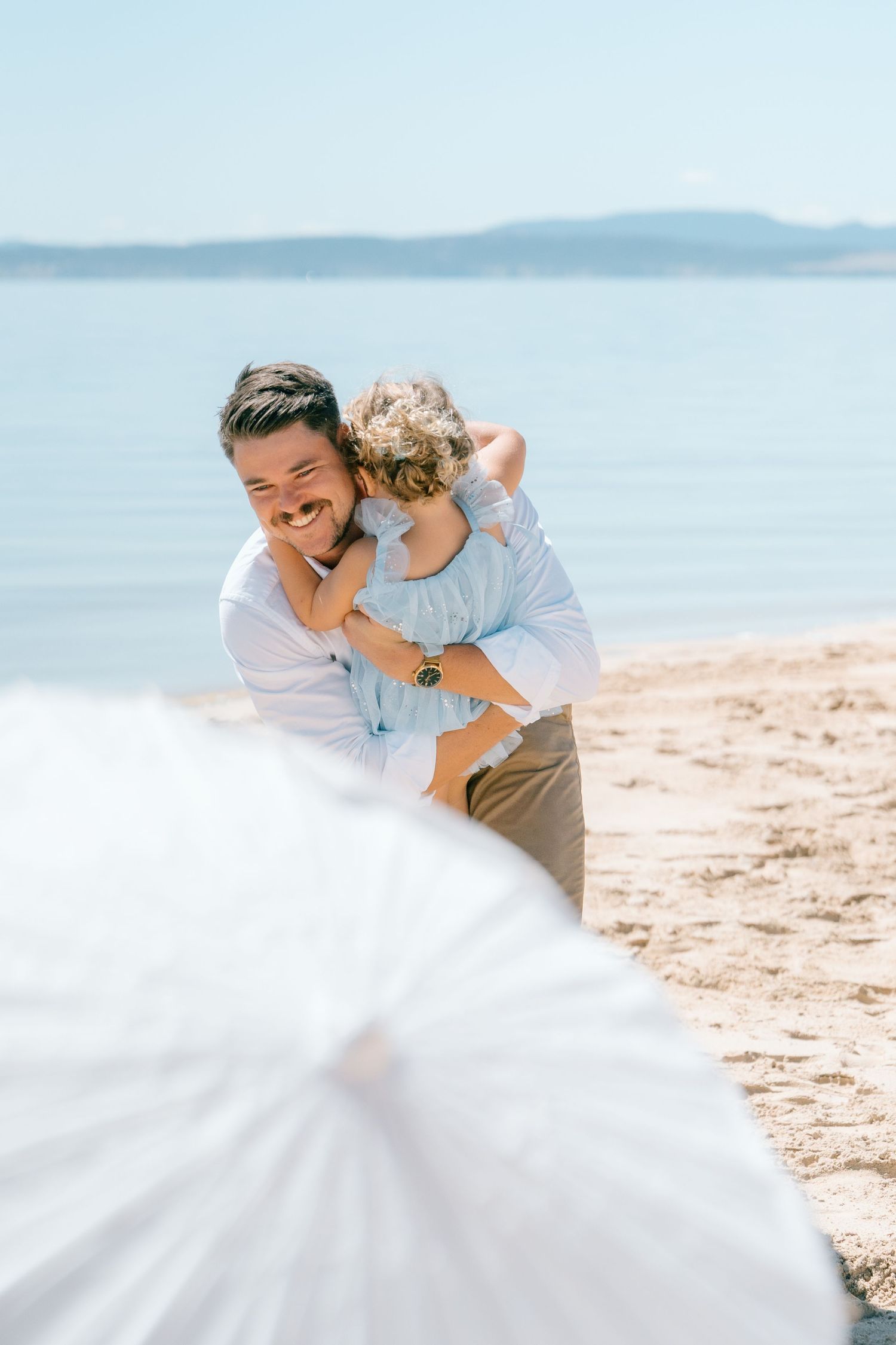 Laughing groom in white shirt hugging curly-haired child on sunny beach with mountains visible across the water.