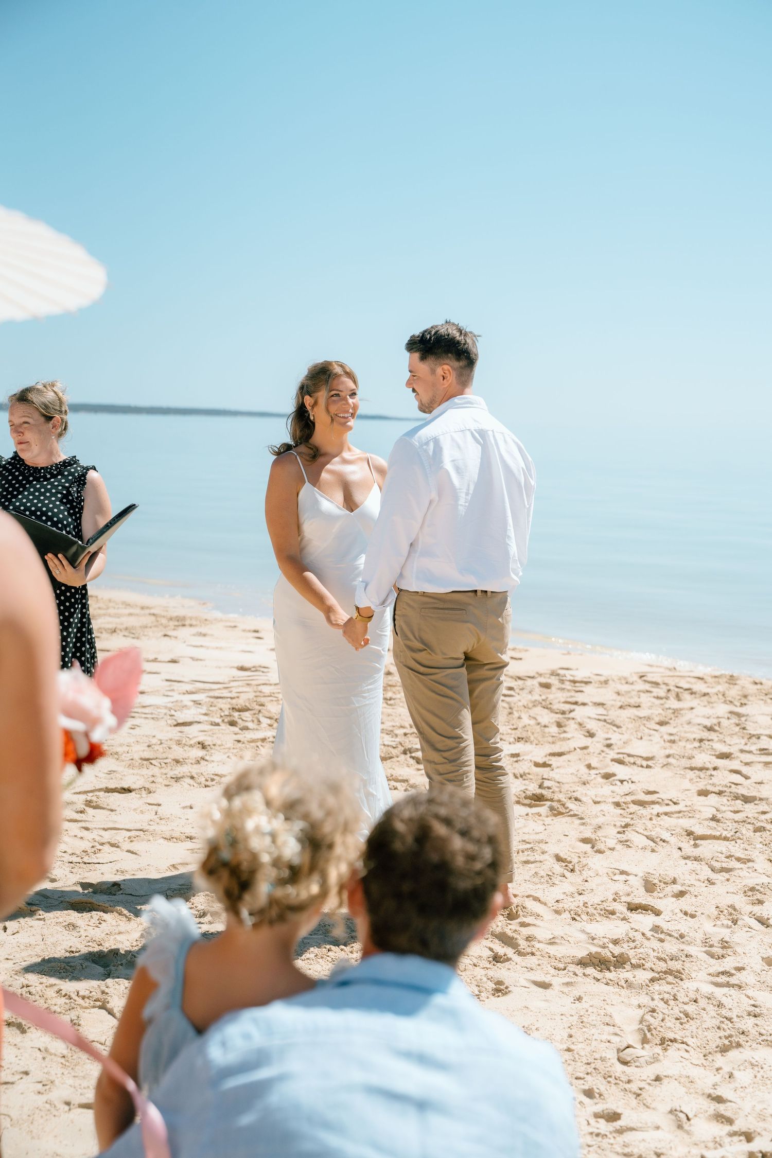 Bride and groom exchanging vows on a sandy beach with guests watching under blue sky.