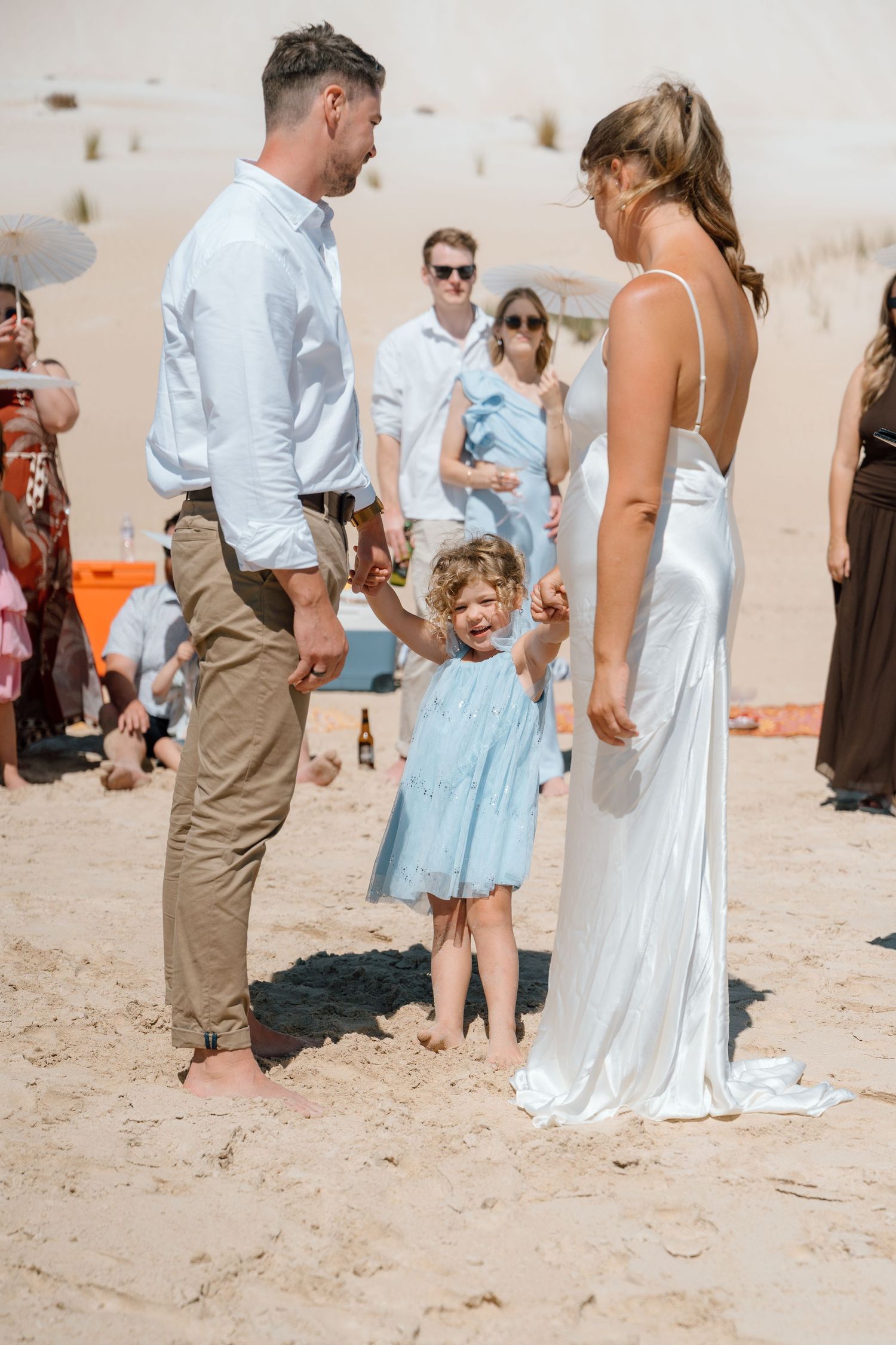Bride and groom holding hands with a young girl in a blue dress during a beach wedding ceremony.
