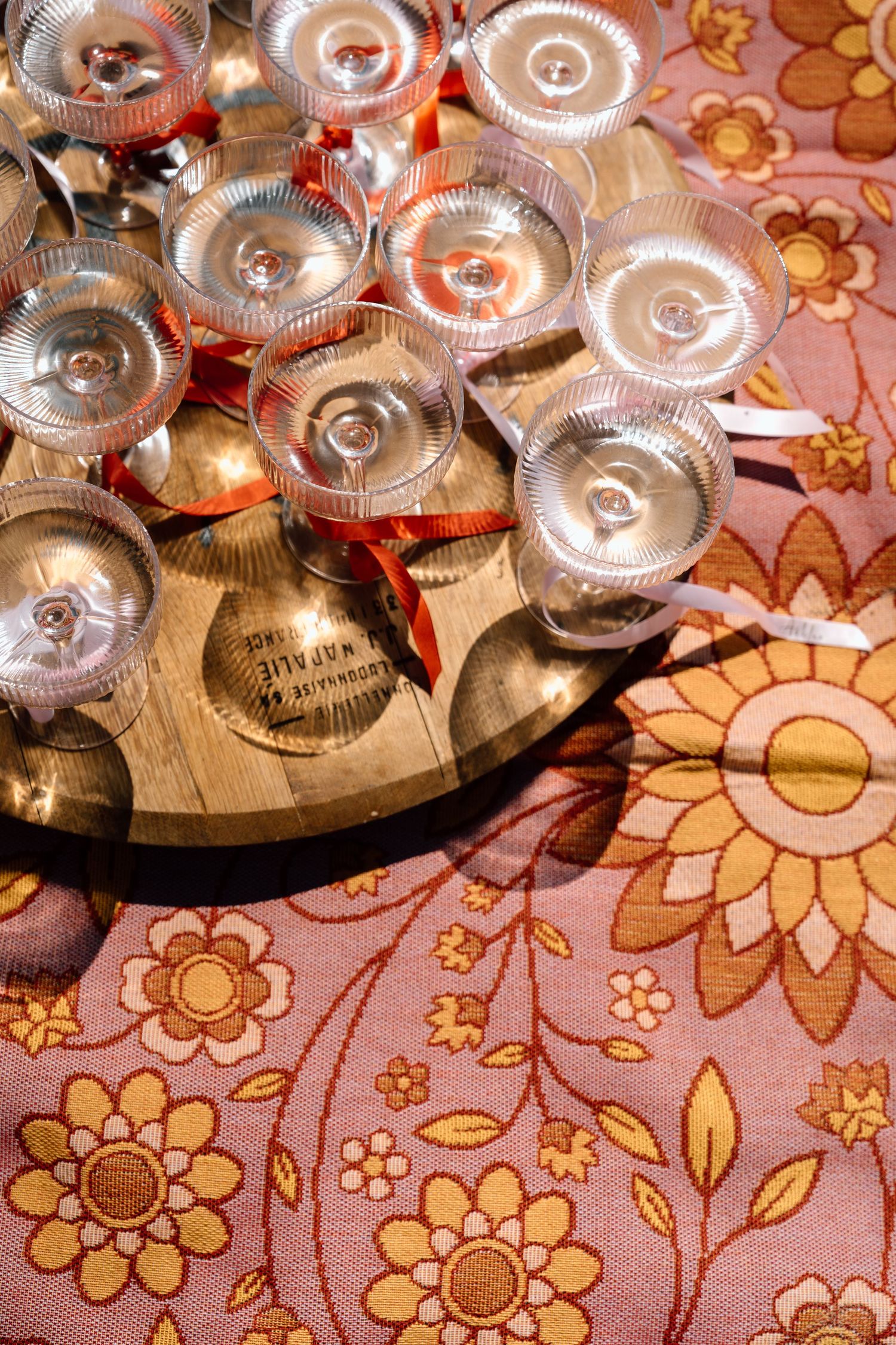 Overhead view of champagne coupe glasses arranged on wooden tray atop floral pink and gold tablecloth.