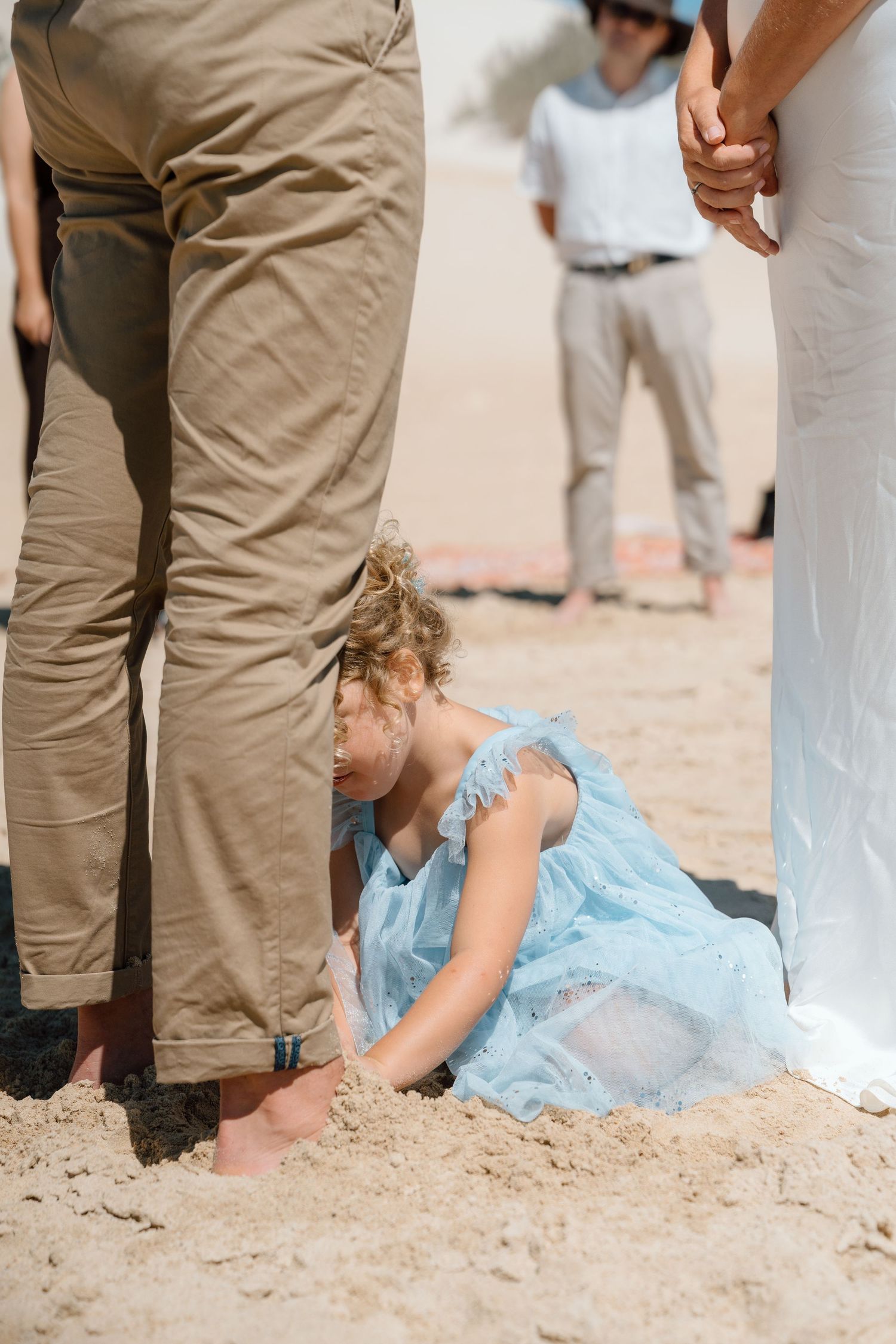 Young girl in blue dress playing in sand between adults at a beach wedding ceremony.
