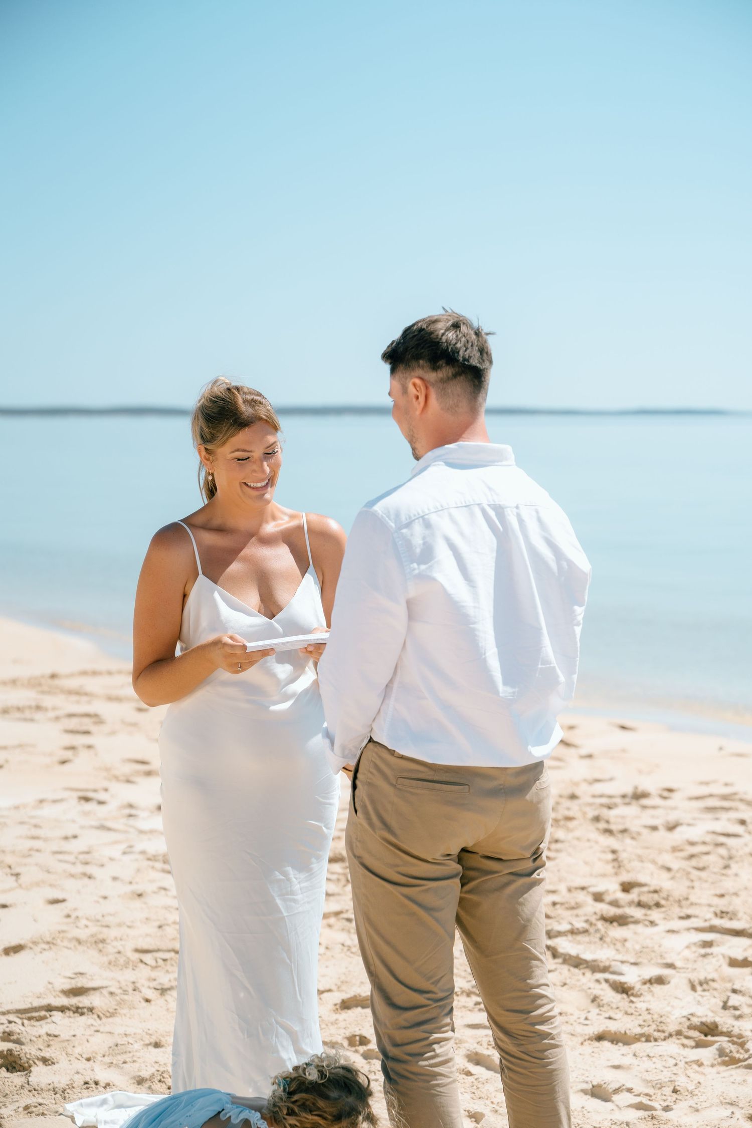 Smiling bride in white slip dress reads vows to groom on sandy beach with calm blue water backdrop.