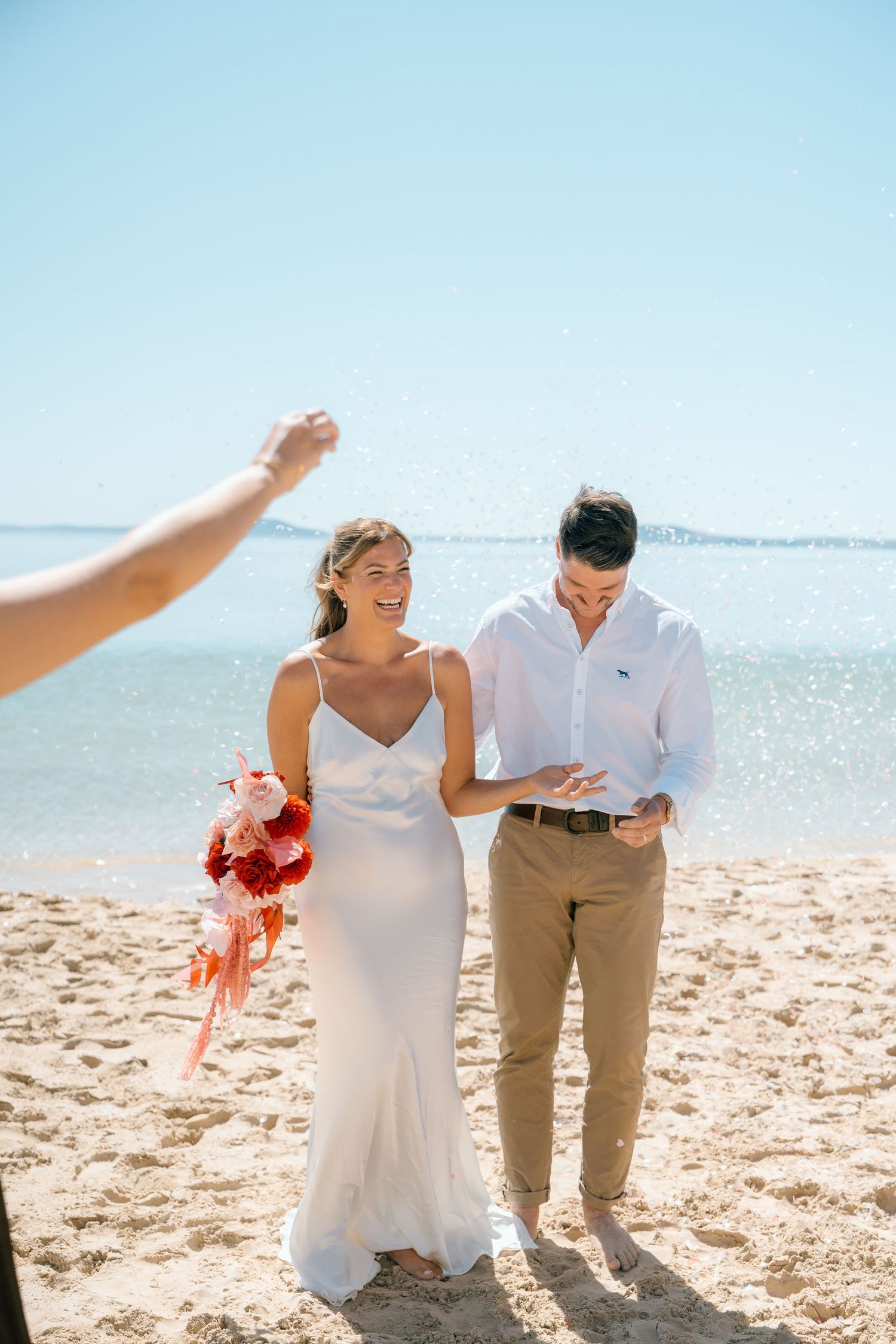 Bride and groom laughing as confetti is thrown at beach wedding ceremony.