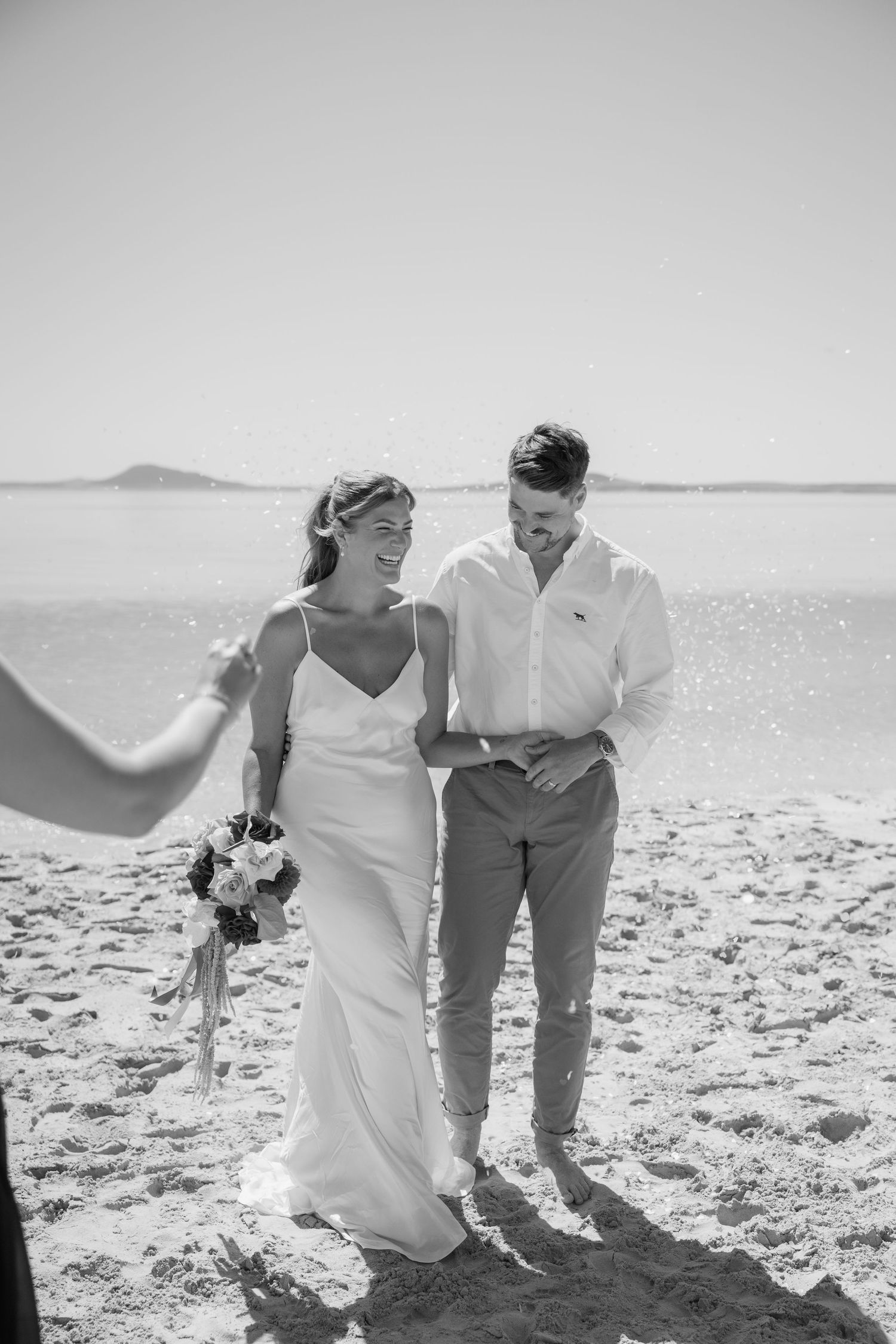 Black and white photo of a smiling bride and groom standing on a beach during their wedding ceremony.