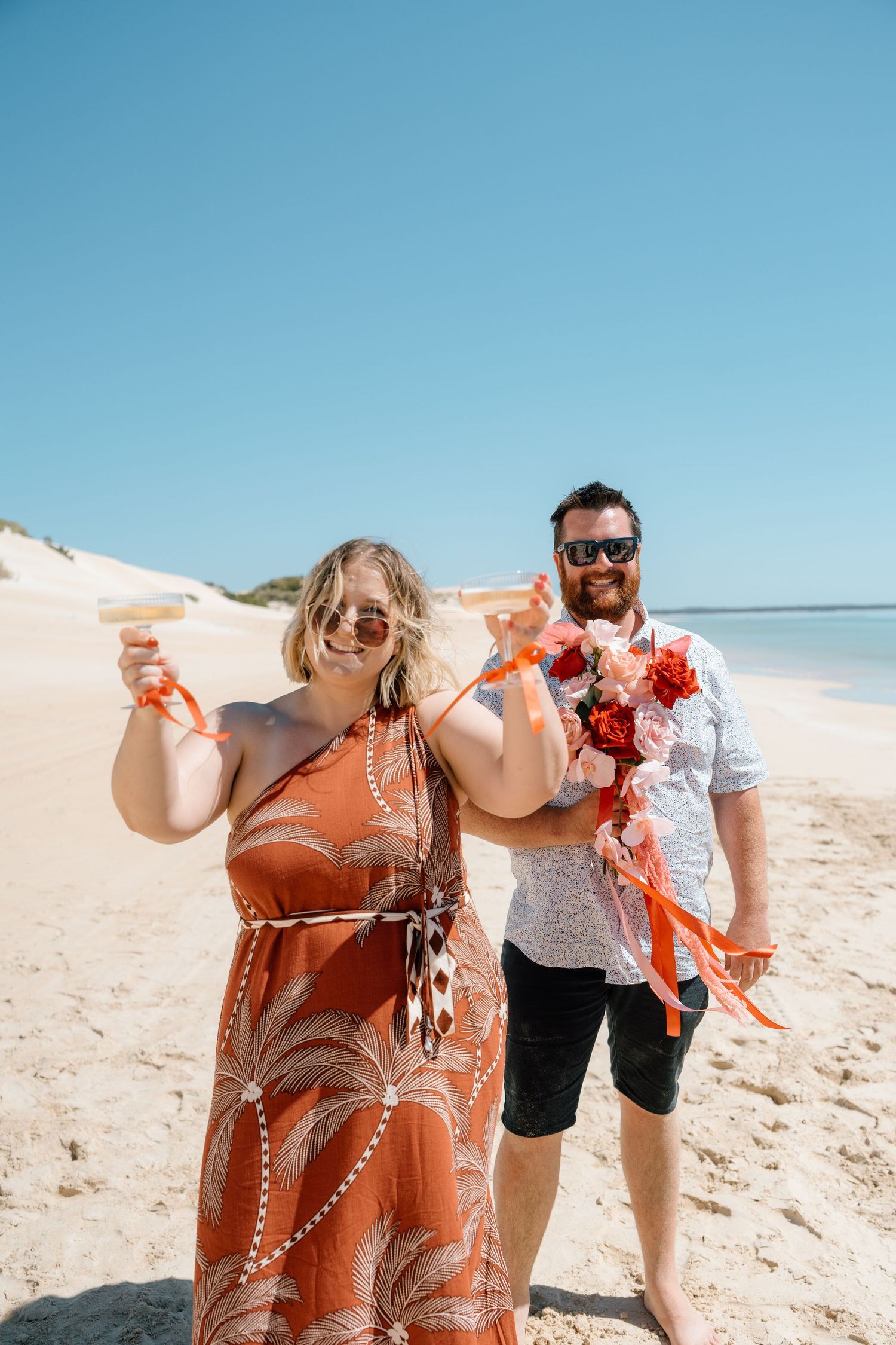 Couple celebrating on a sunny beach, woman in orange dress holding drinks, man in white shirt with floral bouquet.