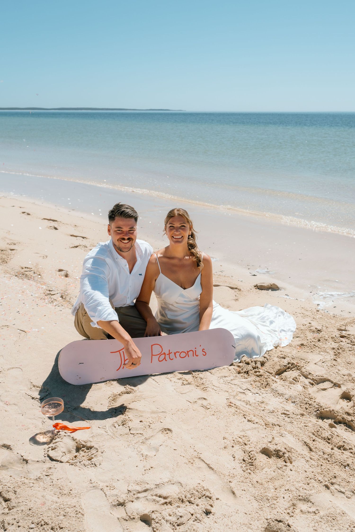Newlywed couple sitting on sandy beach holding a personalized surfboard reading The Patronis with calm ocean behind.