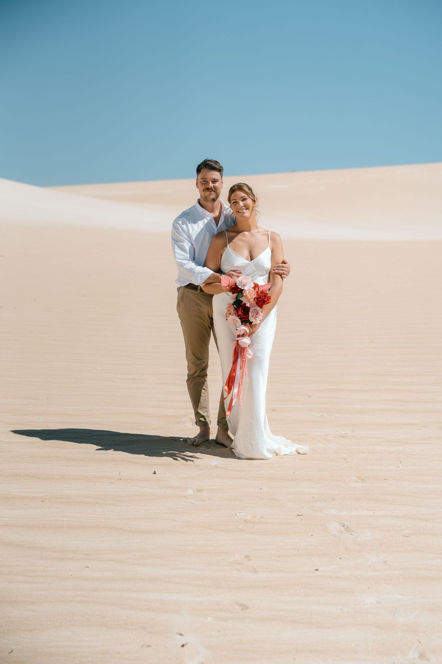 Bride and groom posing together on sandy desert dunes, bride holding red floral bouquet in white gown.