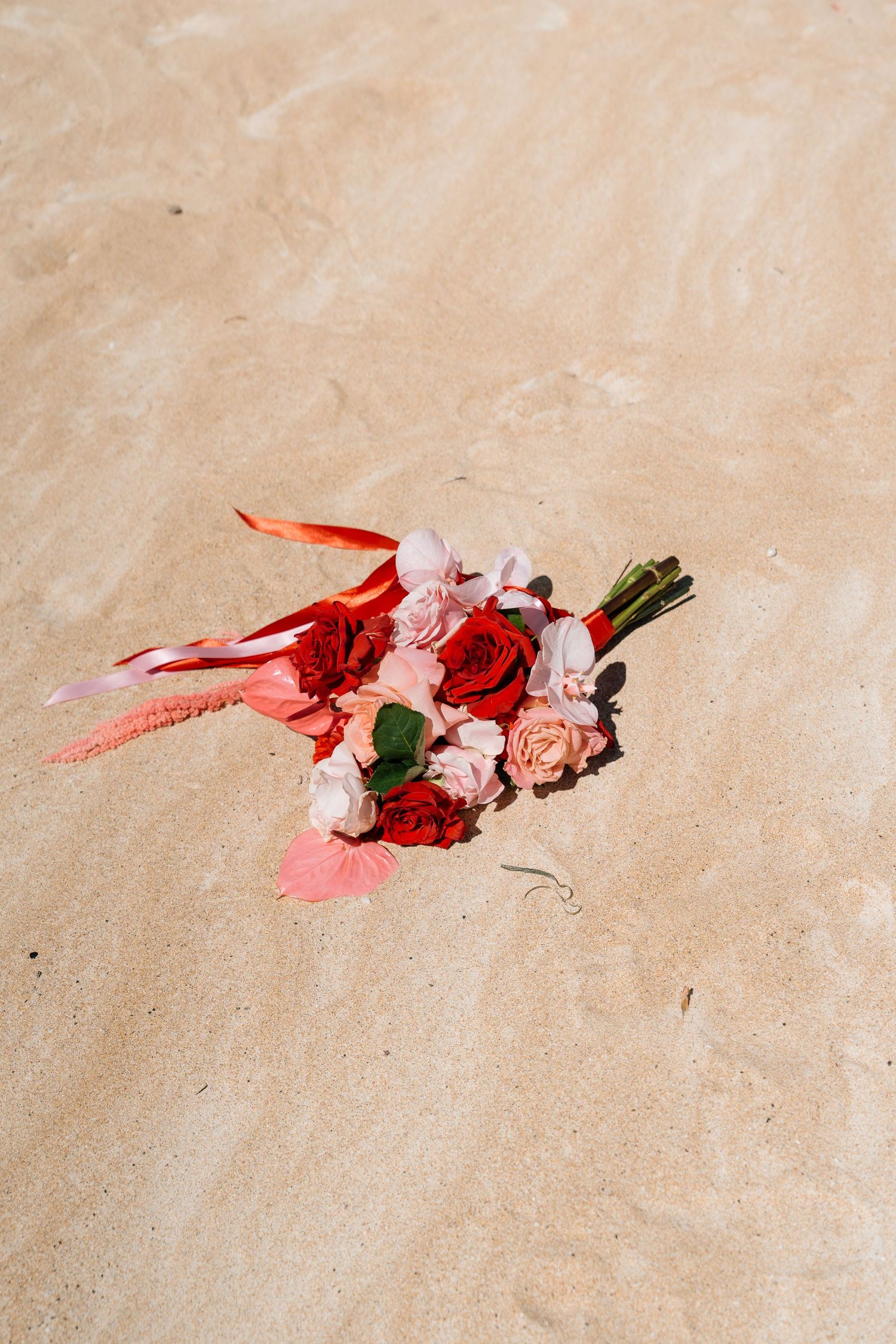 A bridal bouquet of red and pink roses with ribbons resting on sandy beach.