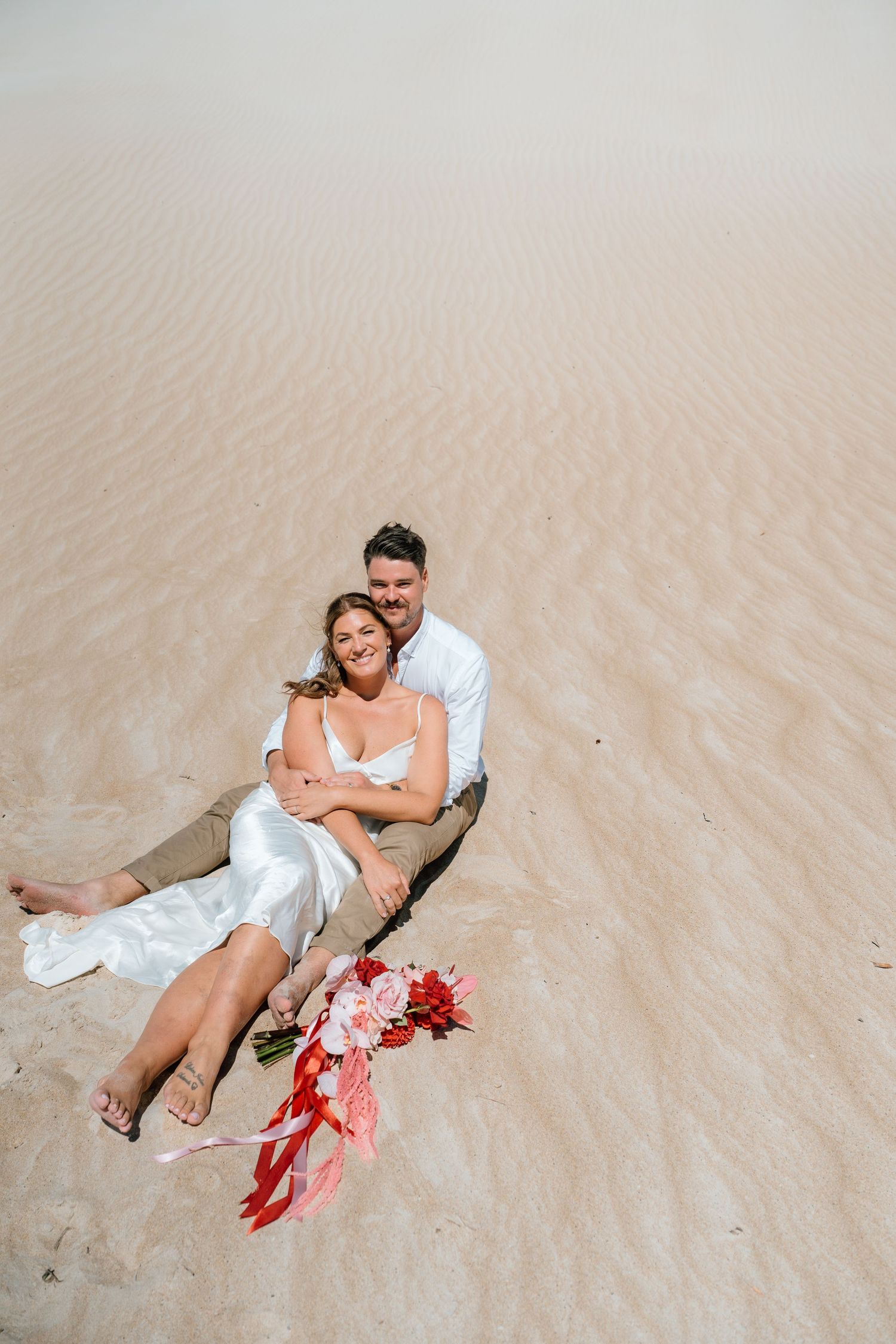 Couple in wedding attire sitting together on sandy dunes, bride holding red bouquet.
