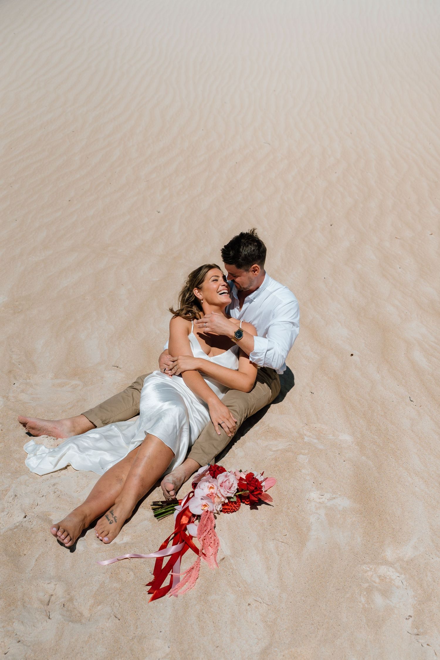 Bride and groom sit together on sandy dunes with red bridal bouquet in wedding photo.
