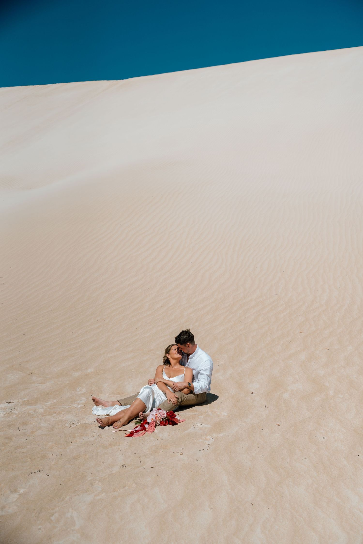 Couple sitting together on sandy desert dunes, embracing romantically with red flowers nearby under a clear blue sky.