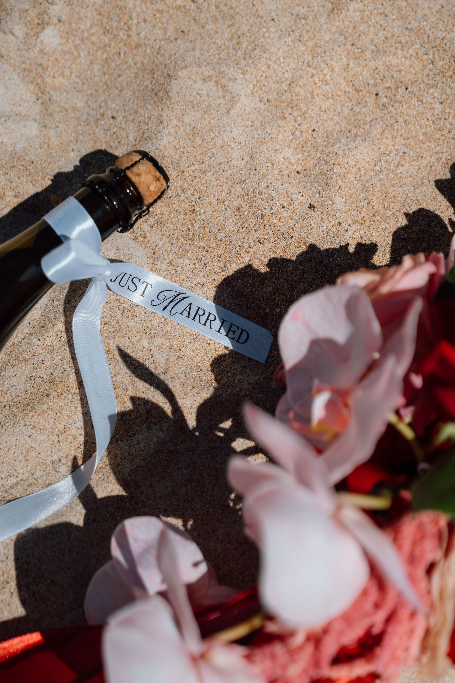 Just Married champagne bottle with ribbon tag lying on sand beside pink orchid wedding flowers.