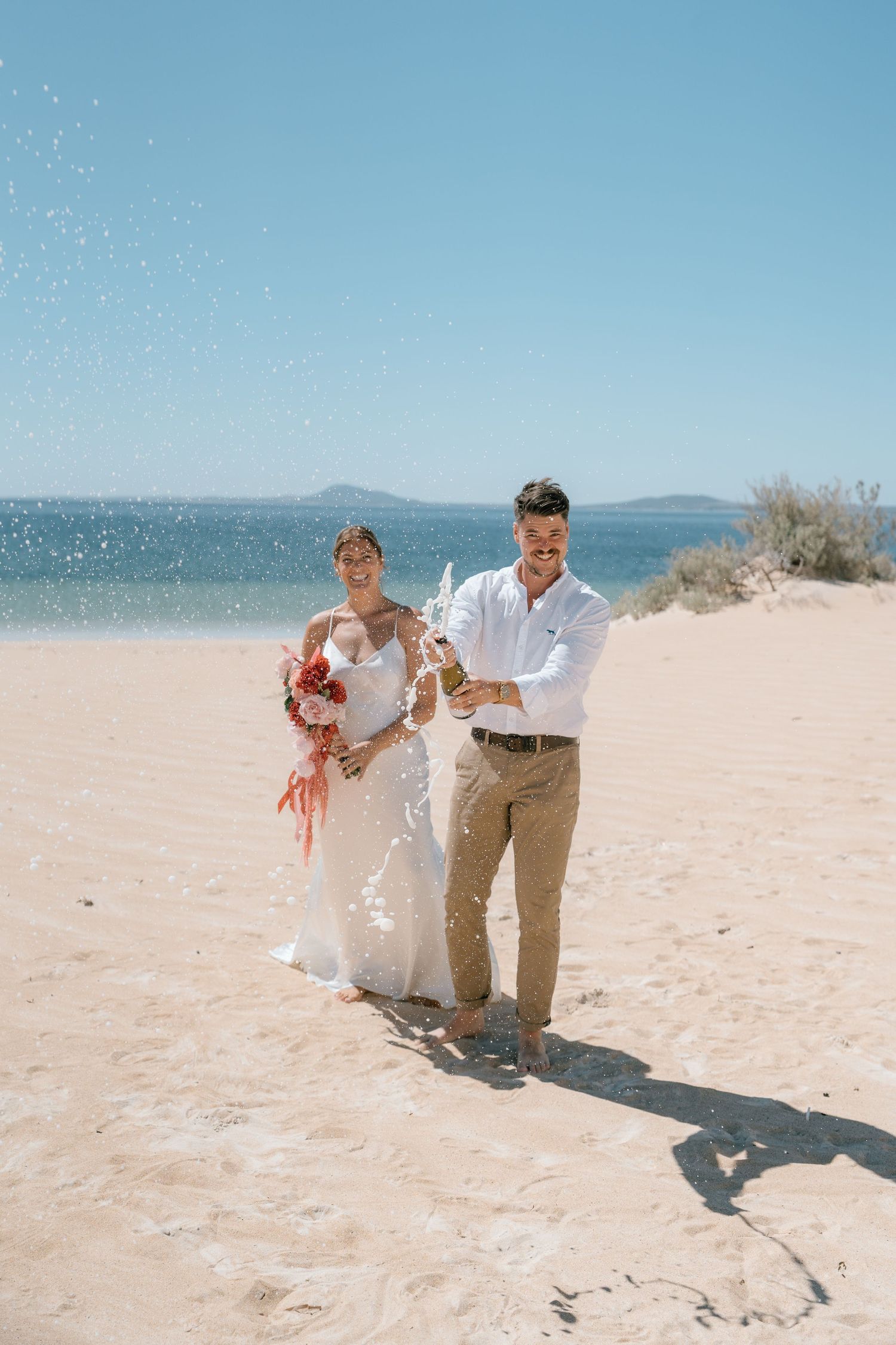 Happy couple celebrating wedding on beach, groom popping champagne bottle with ocean backdrop.
