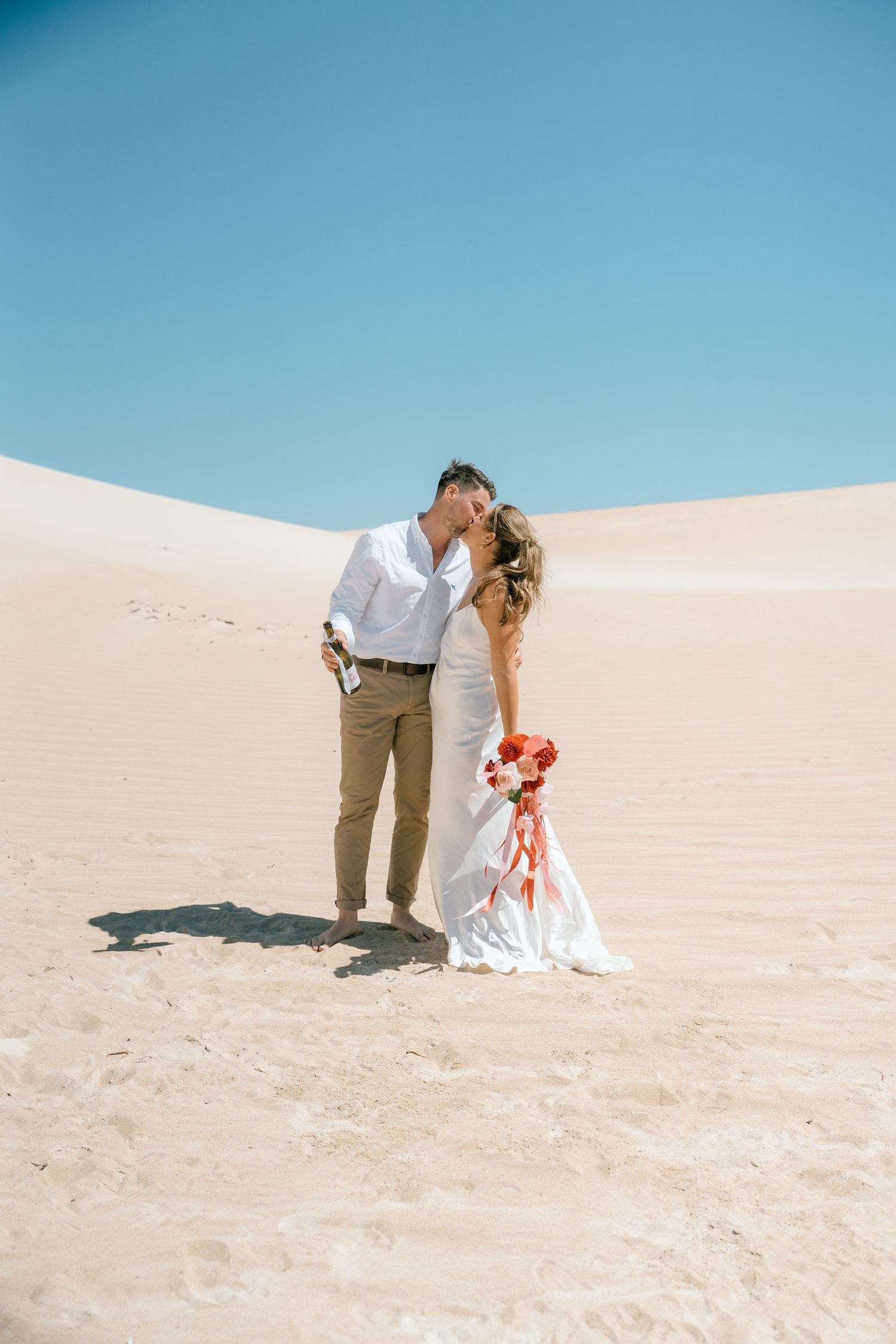 Bride and groom kissing on sandy desert dunes under clear blue sky, bride holding red bouquet.