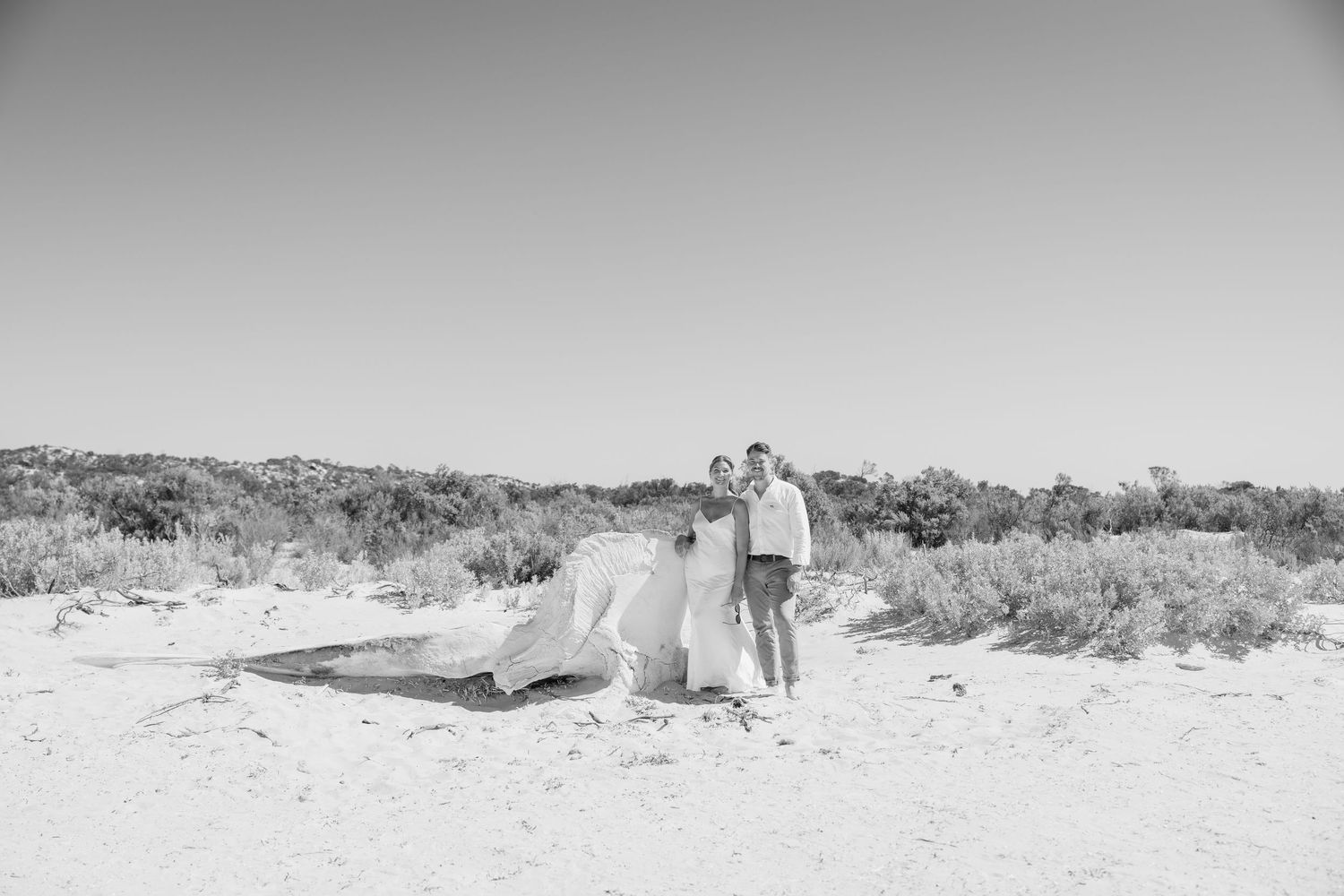 Black and white beach wedding photo of couple standing on white sand with flowing bridal veil and coastal dunes backdrop.