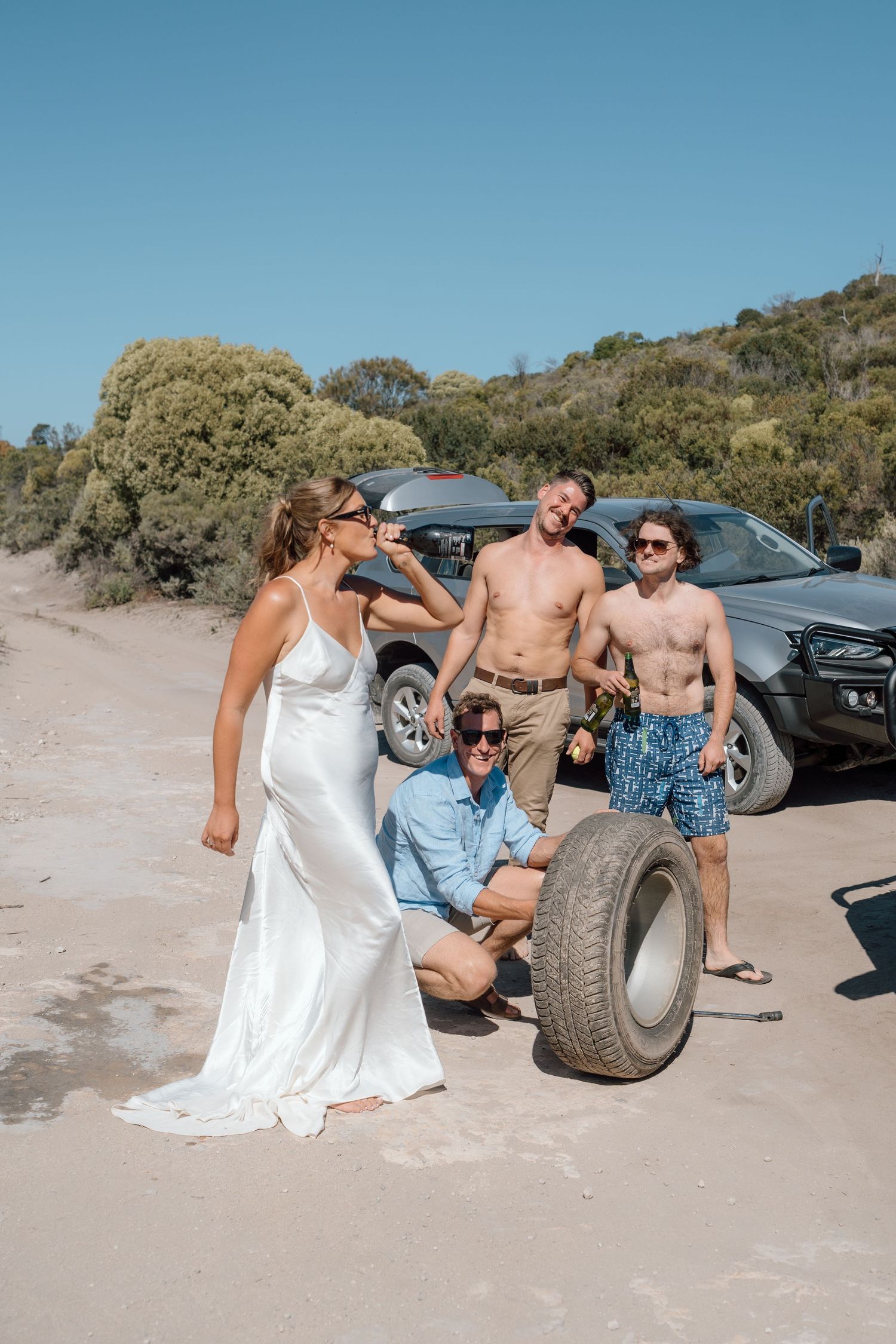 Bride in white slip dress laughing with shirtless groomsmen changing a tire on a sandy beach road.