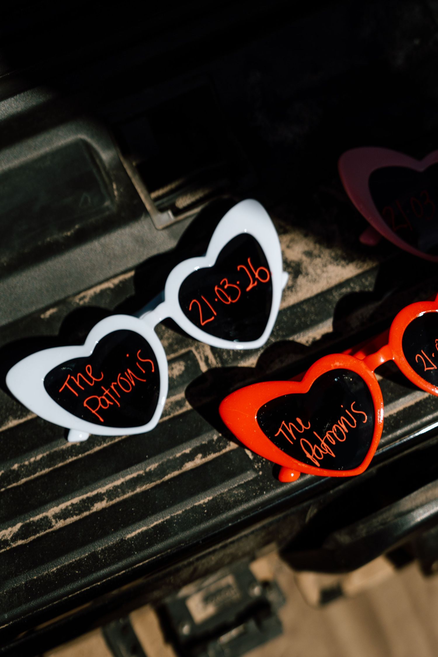 Heart-shaped novelty sunglasses labeled The Patronis with wedding date 21.13.26 displayed on dark vintage surface.