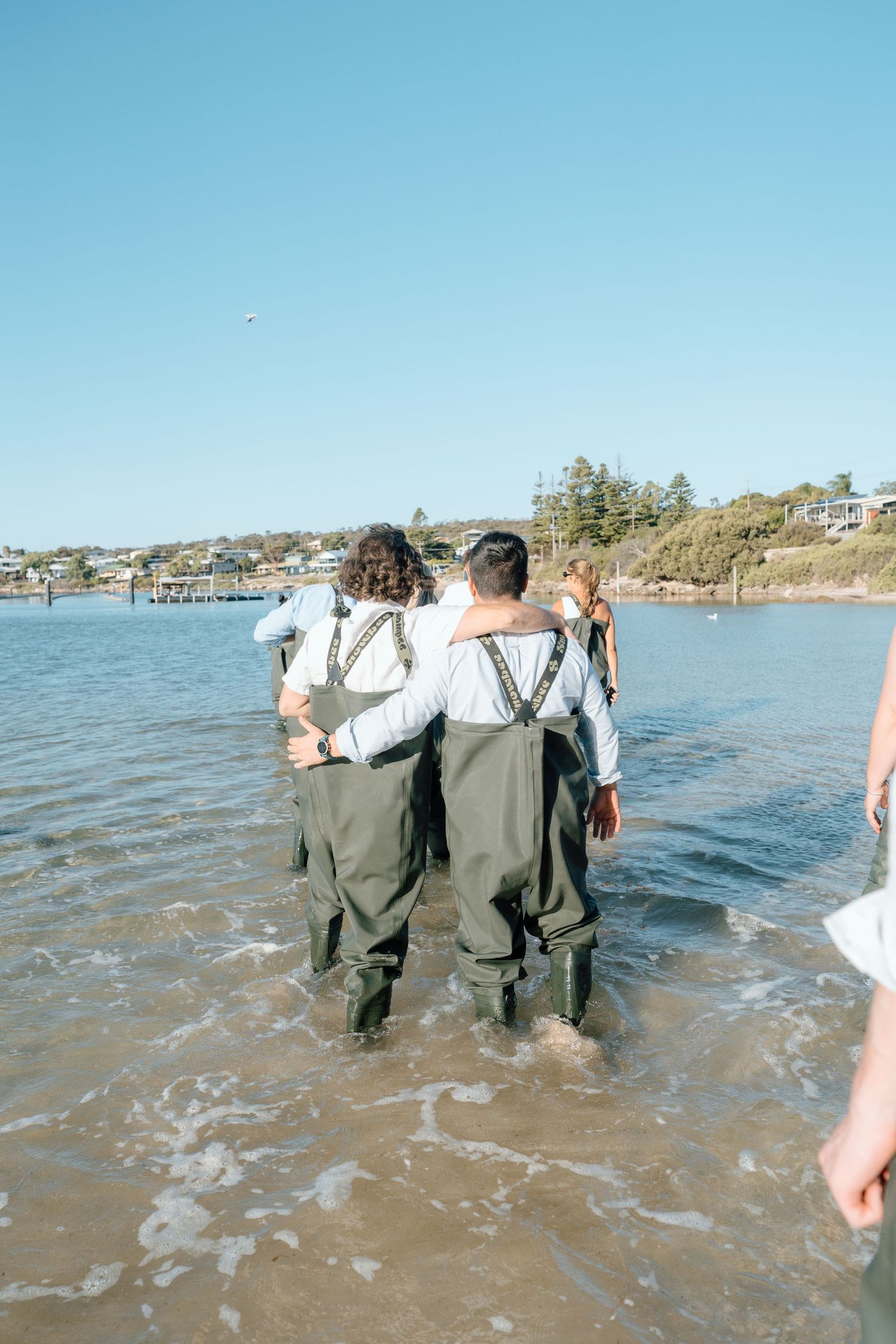 Two groomsmen in waders walk arm-in-arm into ocean water during beach wedding ceremony.