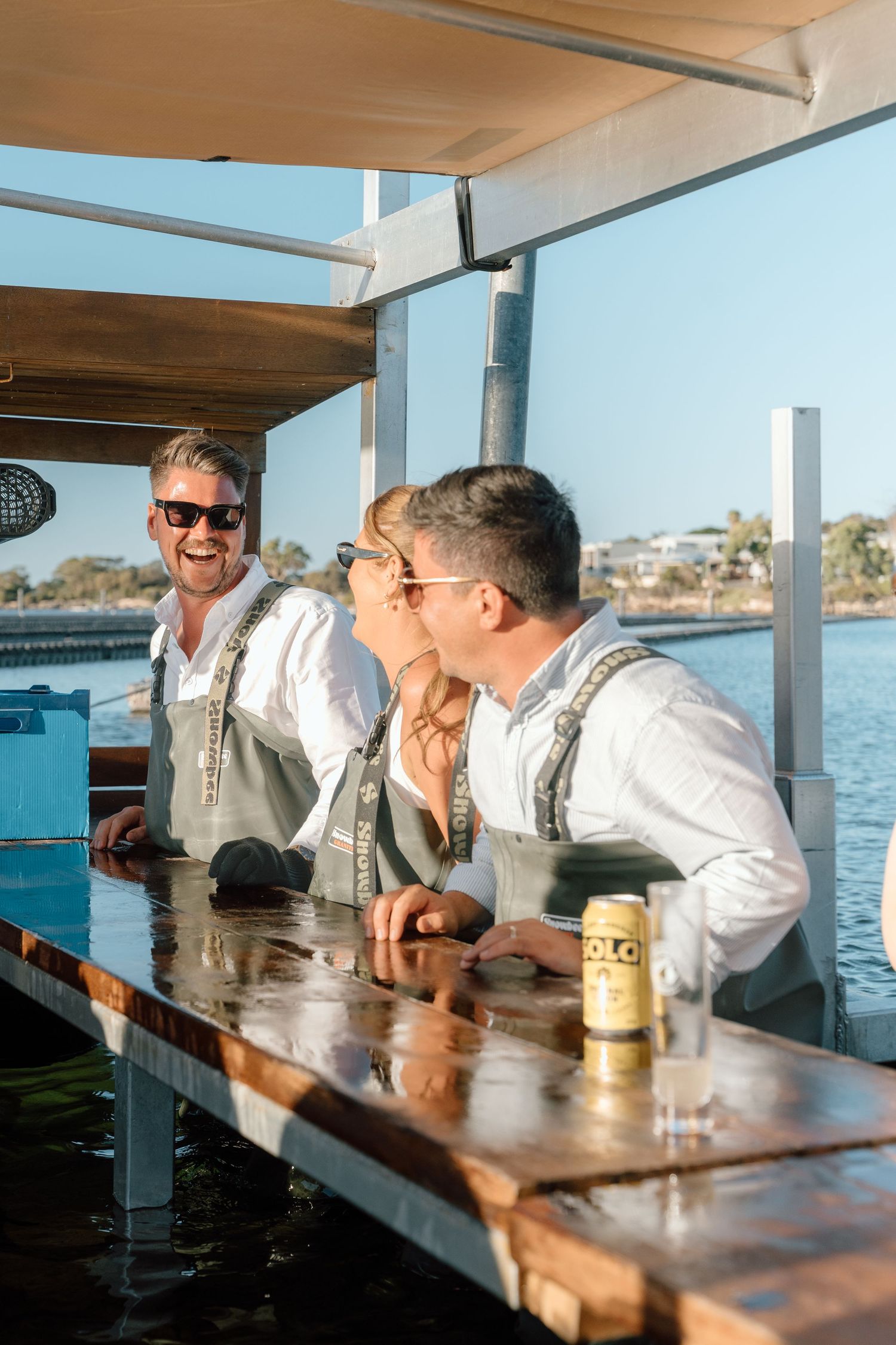 Three friends laughing and enjoying drinks at a waterfront bar on a sunny day.