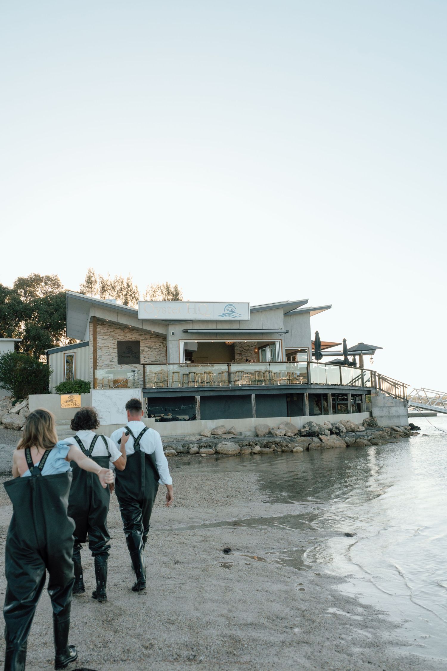 Three people in waders walking along shoreline toward rustic waterfront building at dusk.