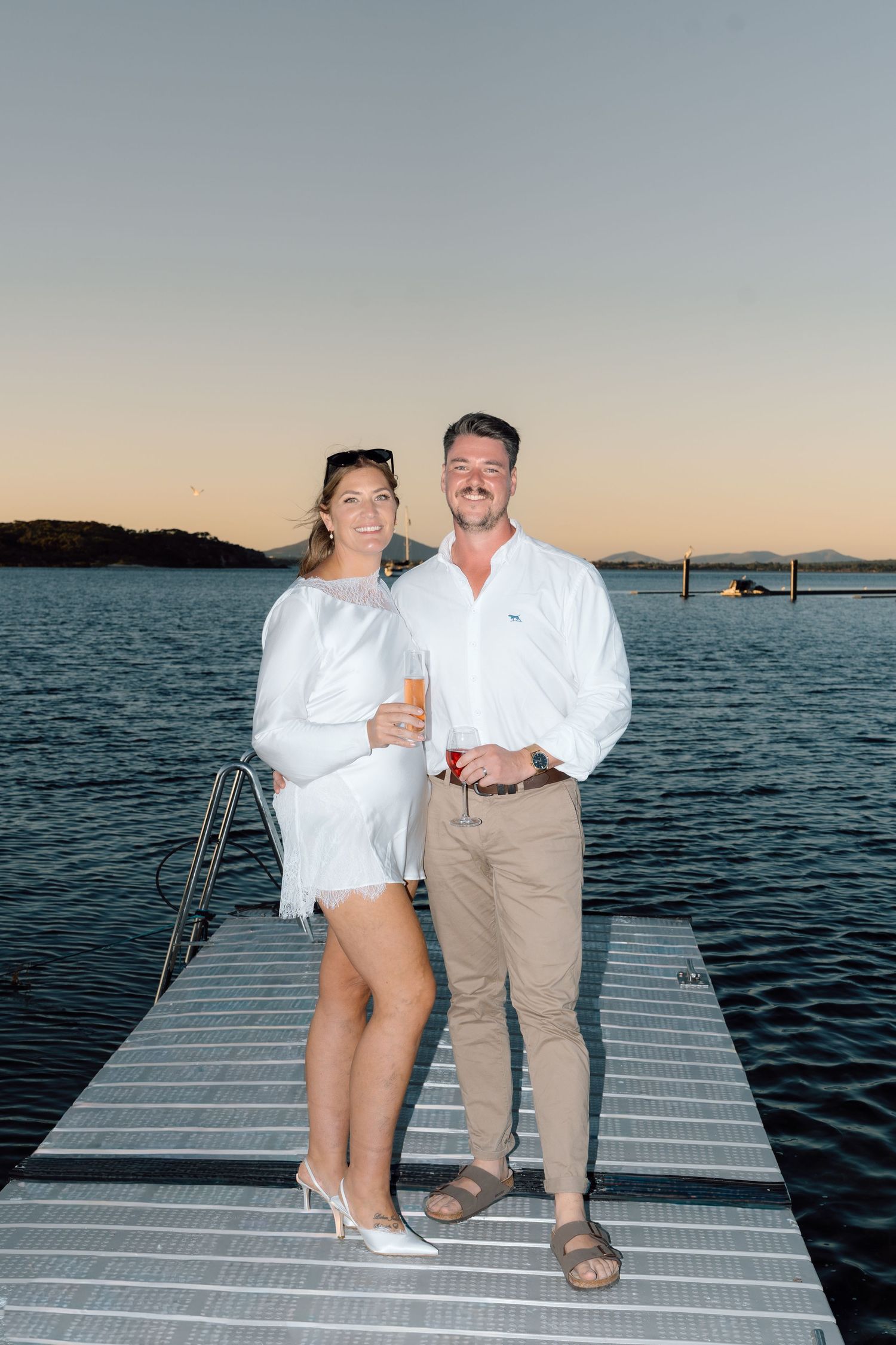 Smiling couple in white outfits holding drinks on a dock at sunset with calm water and hills in background.