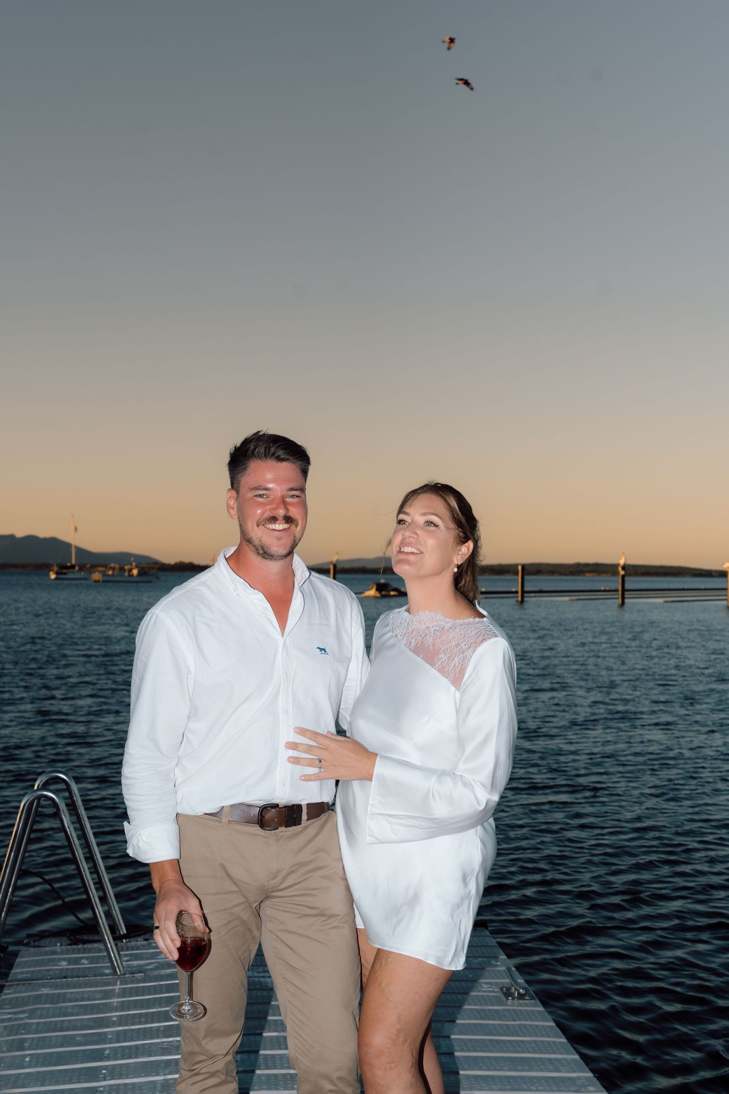 Happy couple in white outfits pose on jetty dock at sunset with wine glass over calm water.